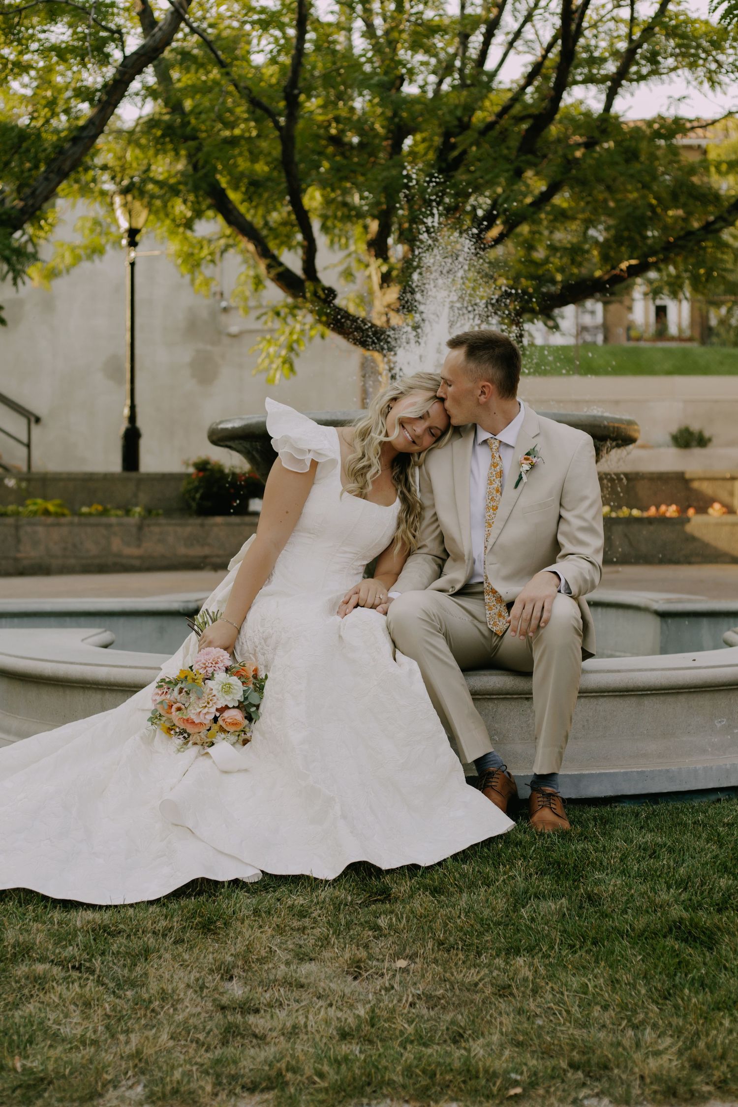 Newlyweds share a romantic moment on grass near a fountain in an outdoor garden setting.