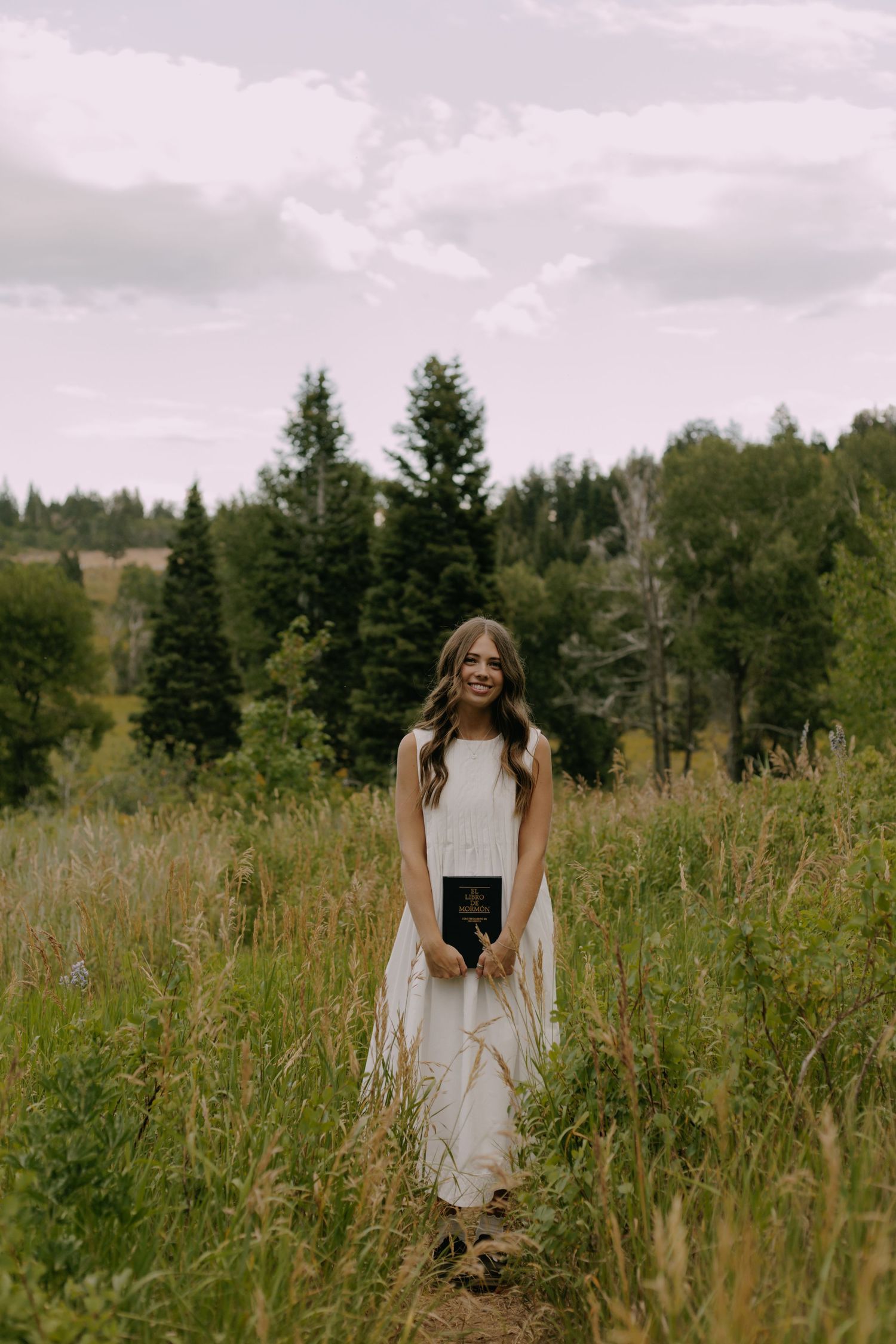 A person in a white dress stands among tall grass with evergreen trees in the background.
