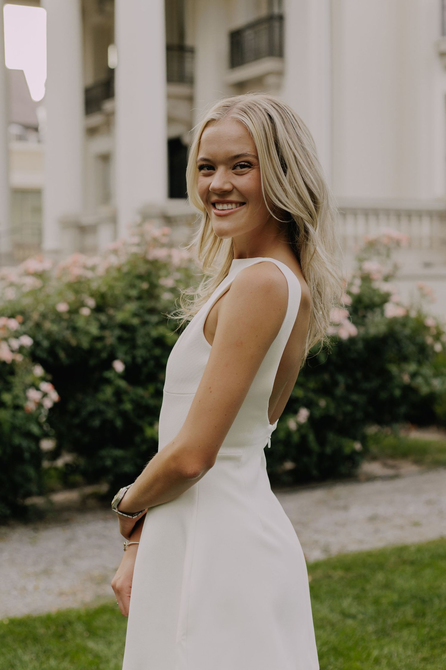 Graduate in white dress poses near rose bushes and classical architecture.