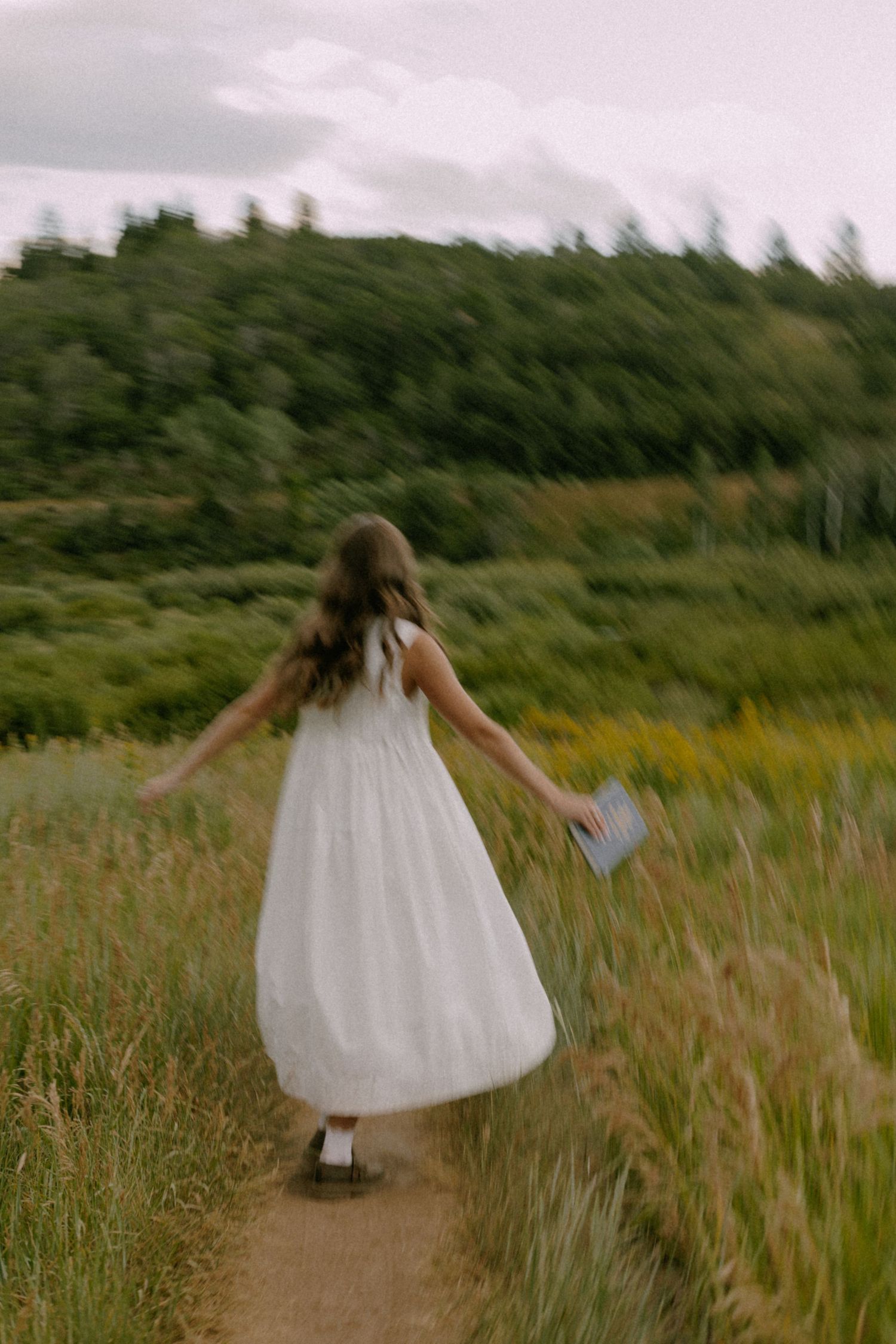 A figure in a flowing white dress twirls through a grassy meadow on a summer day.