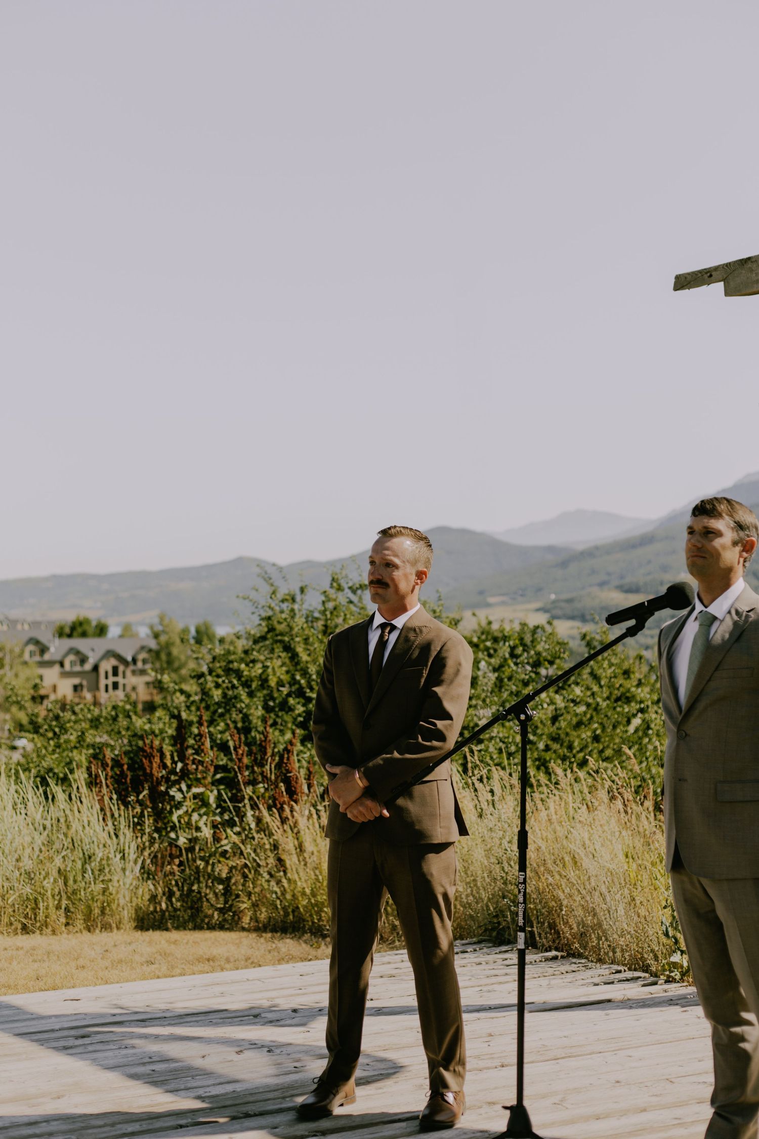 Outdoor wedding ceremony setup with mountain views and men in suits.