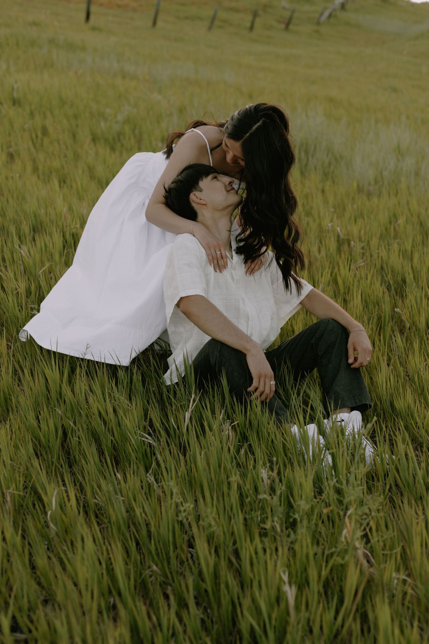 Two people in white clothing embrace while lying in tall grass outdoors.