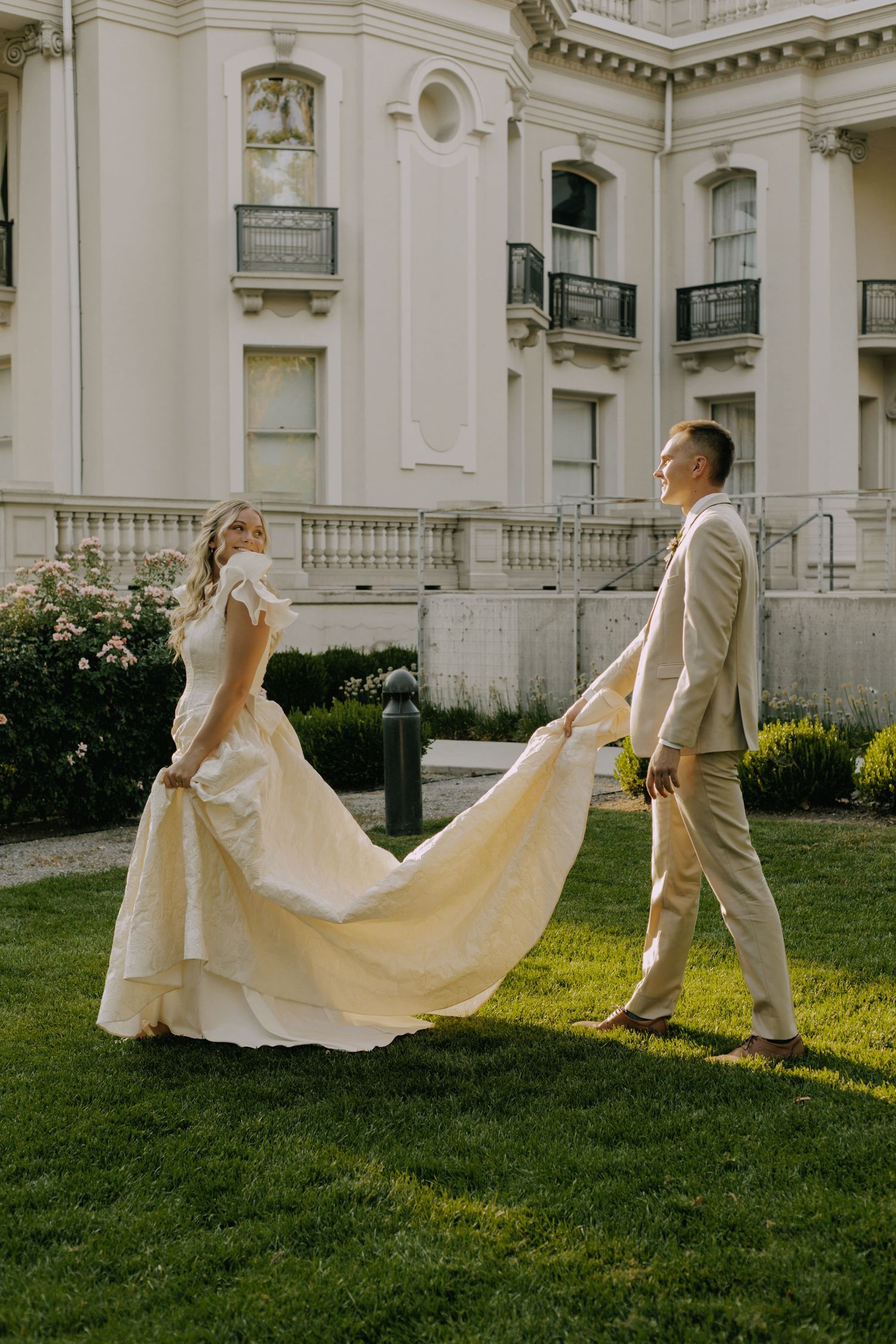 Playful wedding couple spins and dances with dress on lawn by classical building.