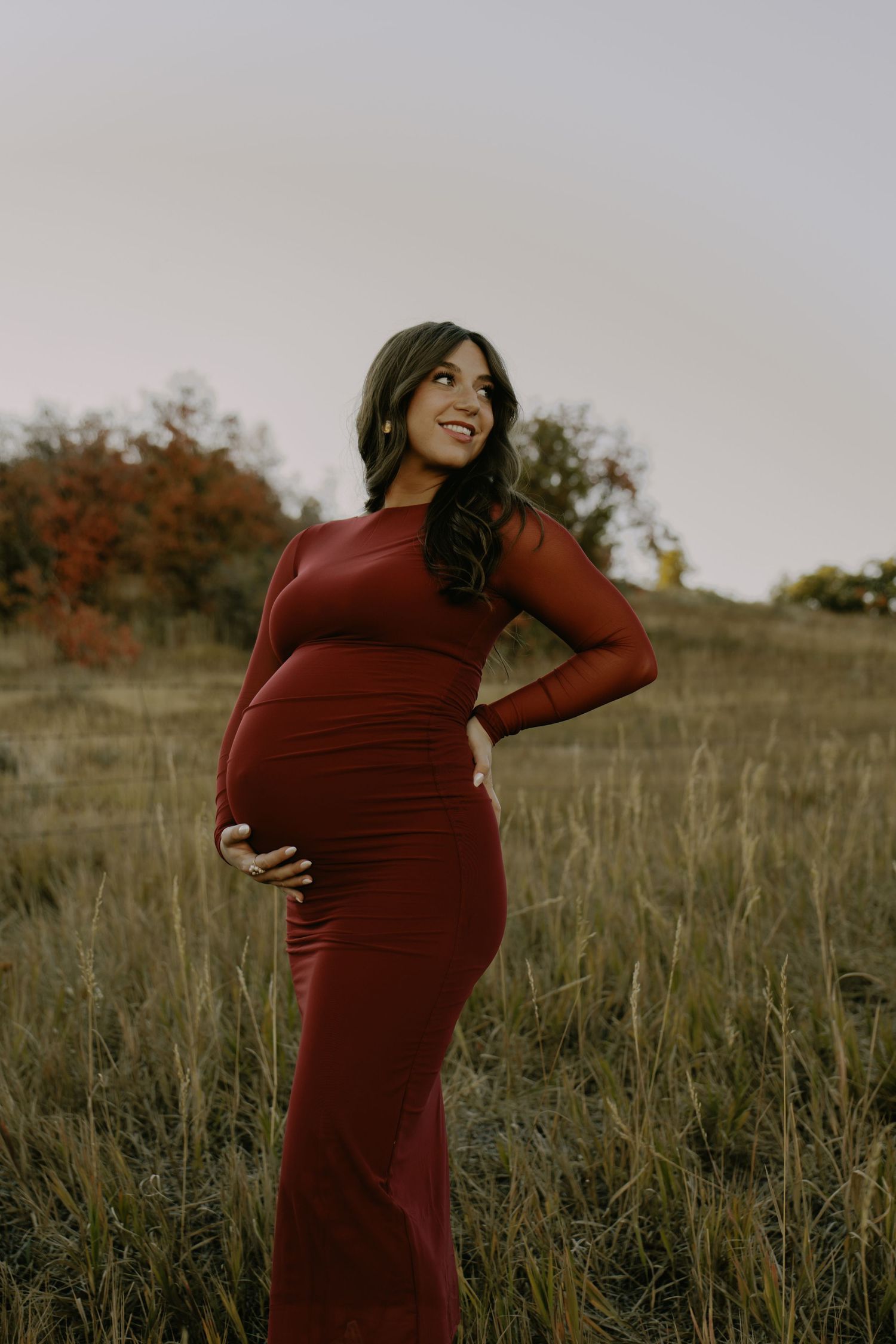 Woman in burgundy dress poses in golden field during maternity photoshoot at sunset.
