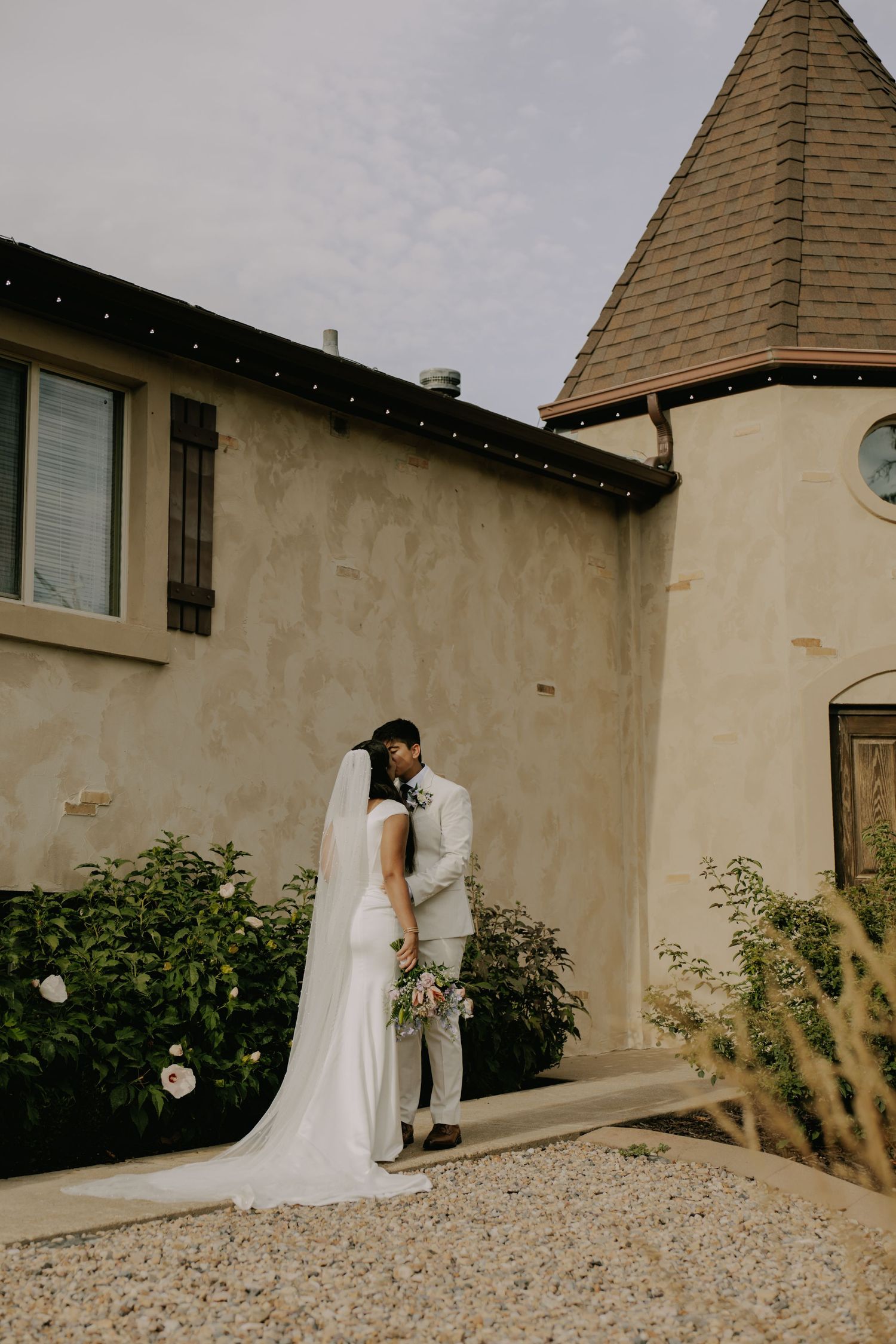 A romantic wedding scene unfolds against a beige stone building with a turret.