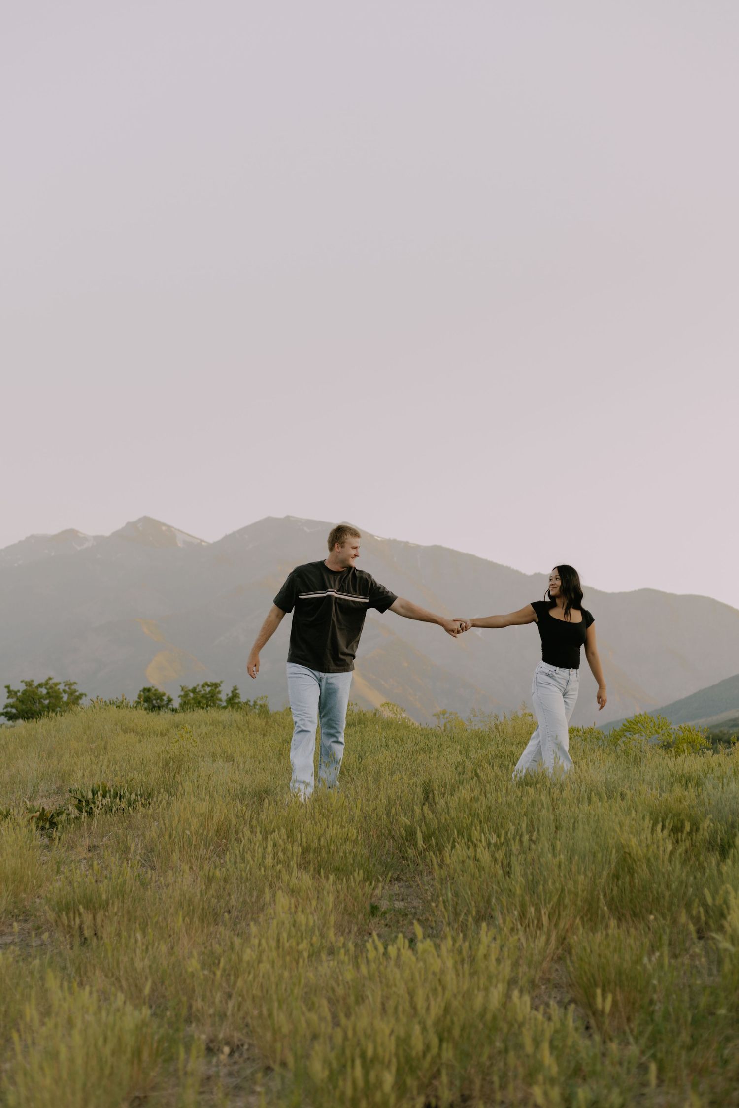 A couple playfully holds hands while standing in a grassy field with mountains in the background at sunset.