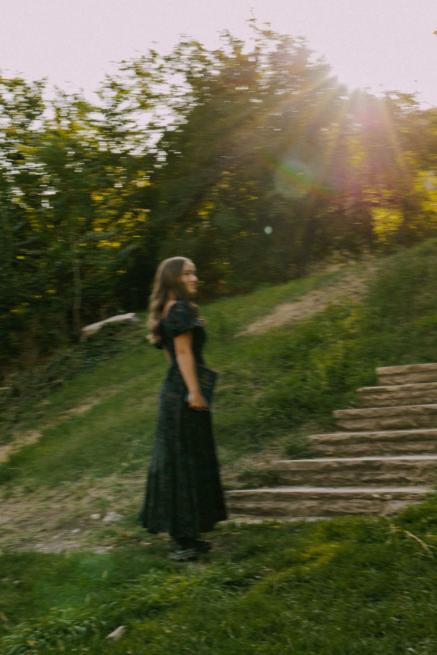 Person in black dress walks up sunlit stone steps surrounded by greenery.