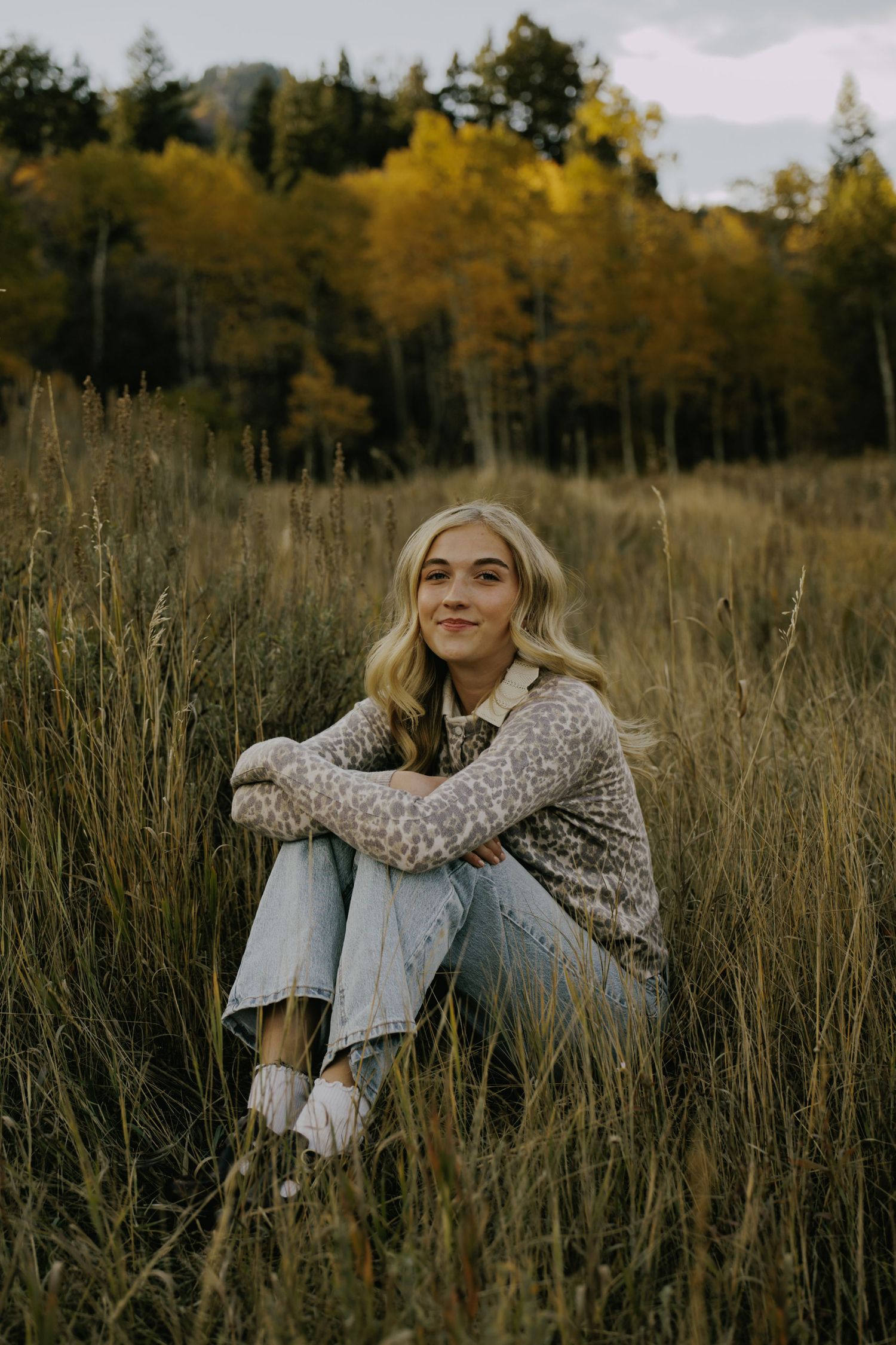 Woman in cozy sweater sits in tall autumn grass during golden hour.