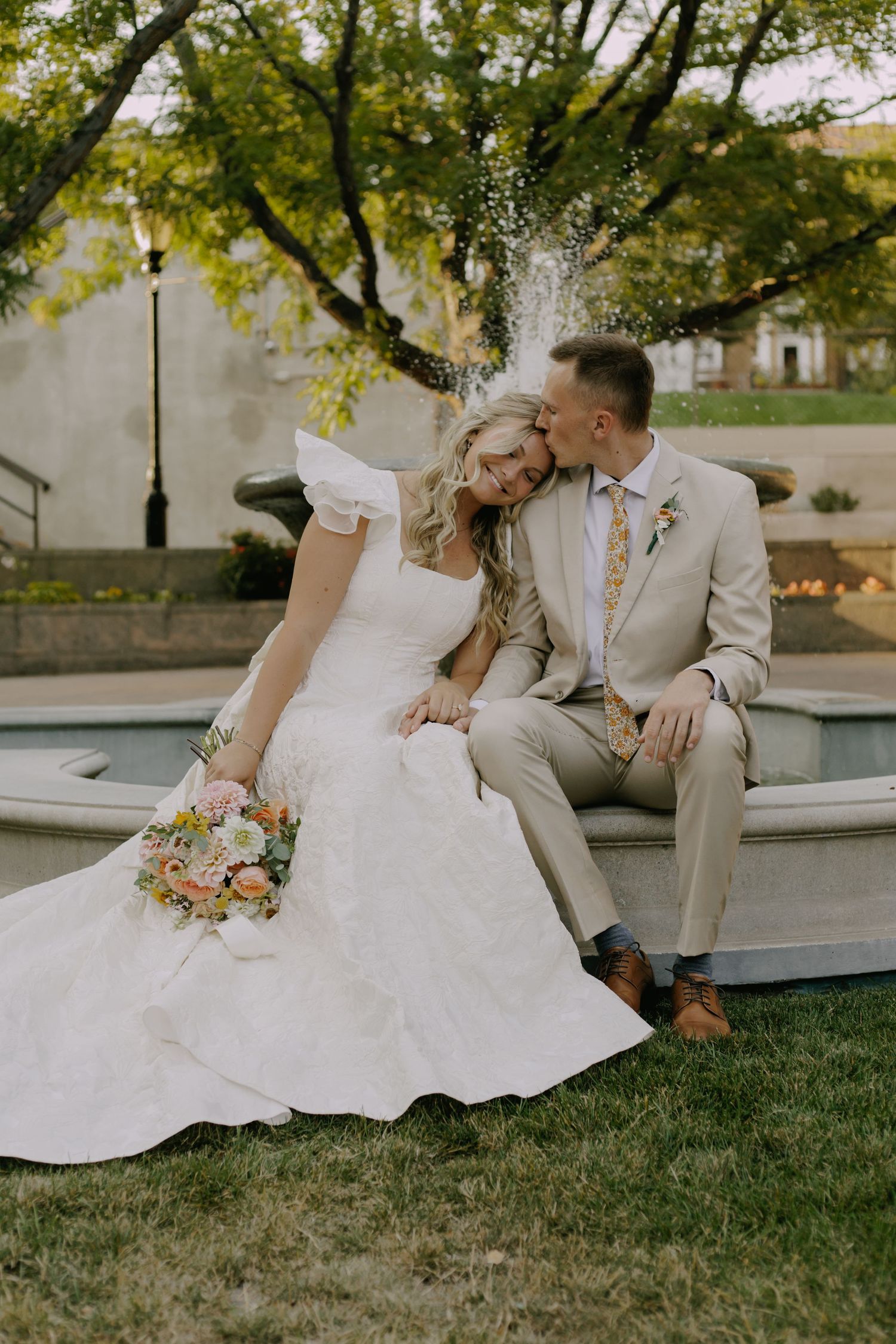 Bride in white dress and groom in tan suit share intimate moment on grassy lawn.