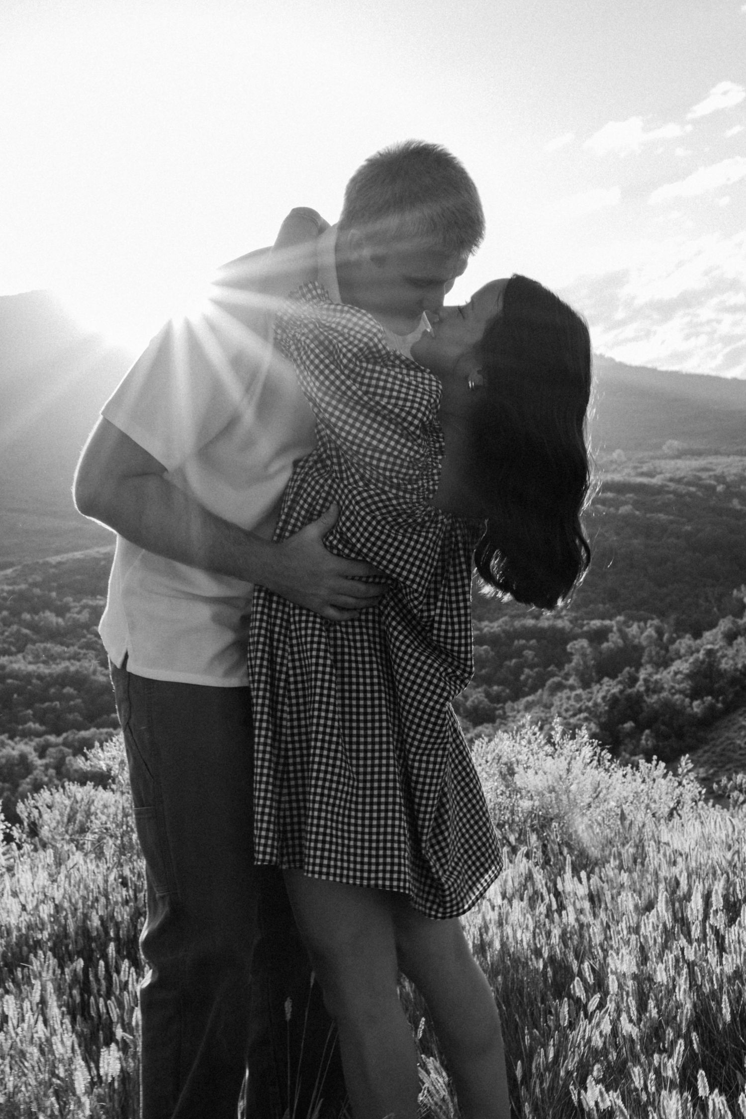 A couple embracing in dramatic sunlight with mountain views and tall grass in the background.