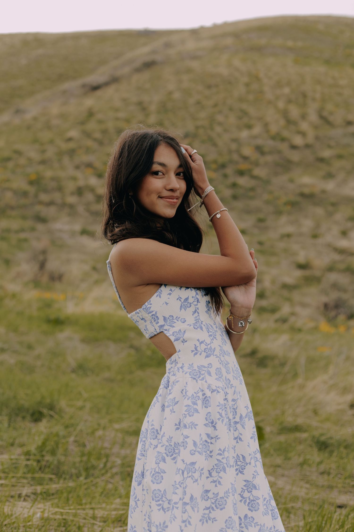 A person poses in a blue and white floral dress against a natural hillside backdrop.