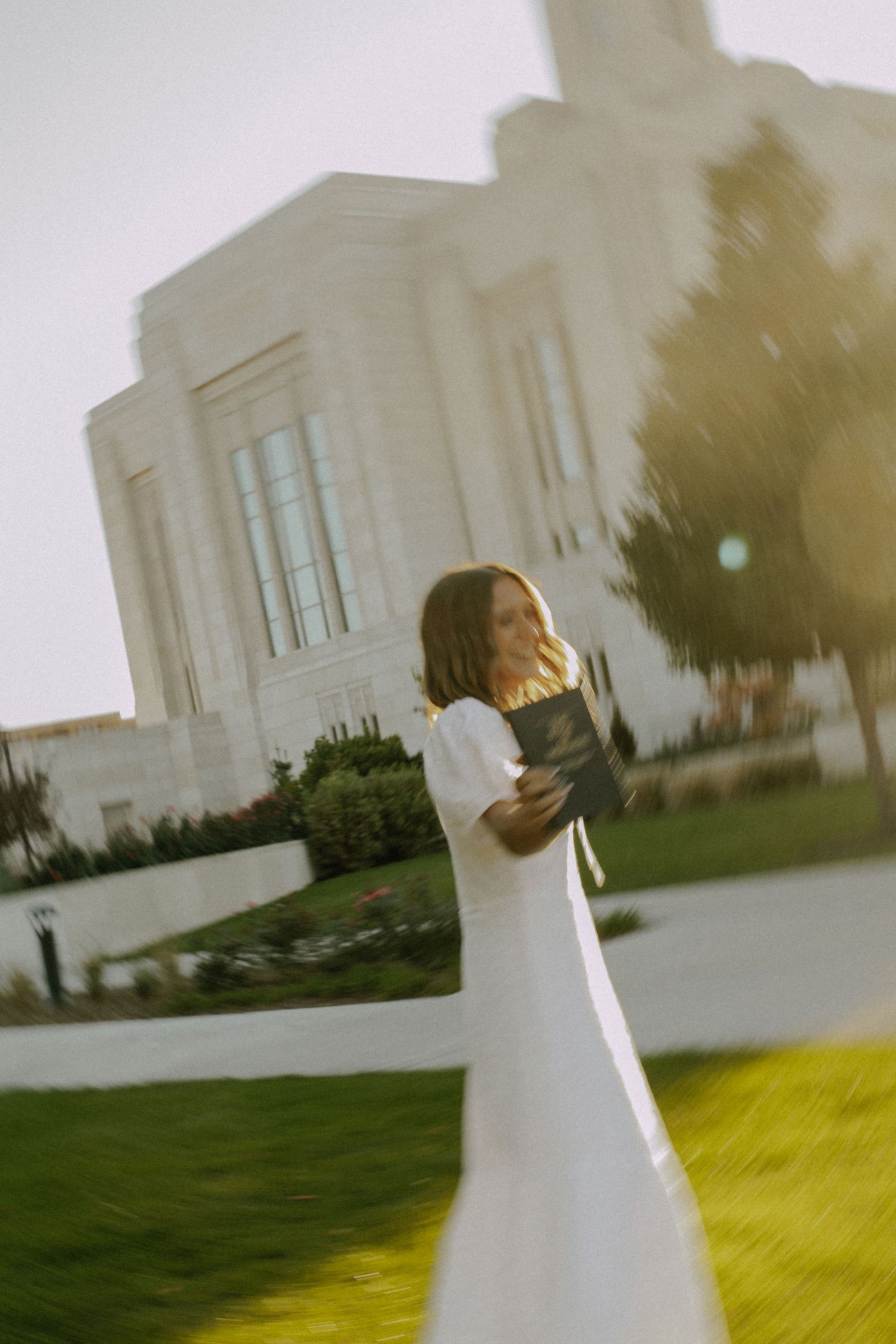 Bride twirls in white dress on sunny lawn with building in background.