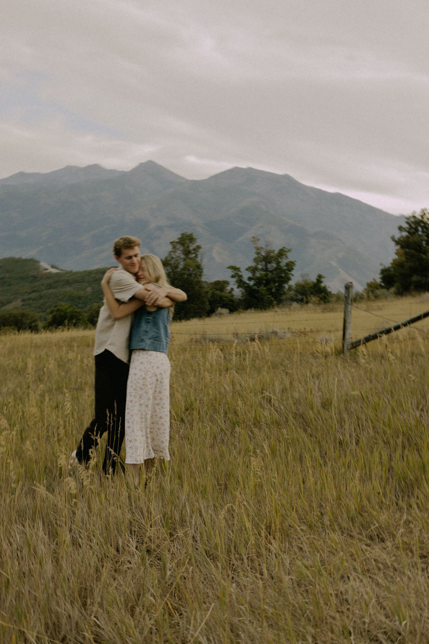A romantic couple embraces in a mountain meadow with dramatic peaks in the background.