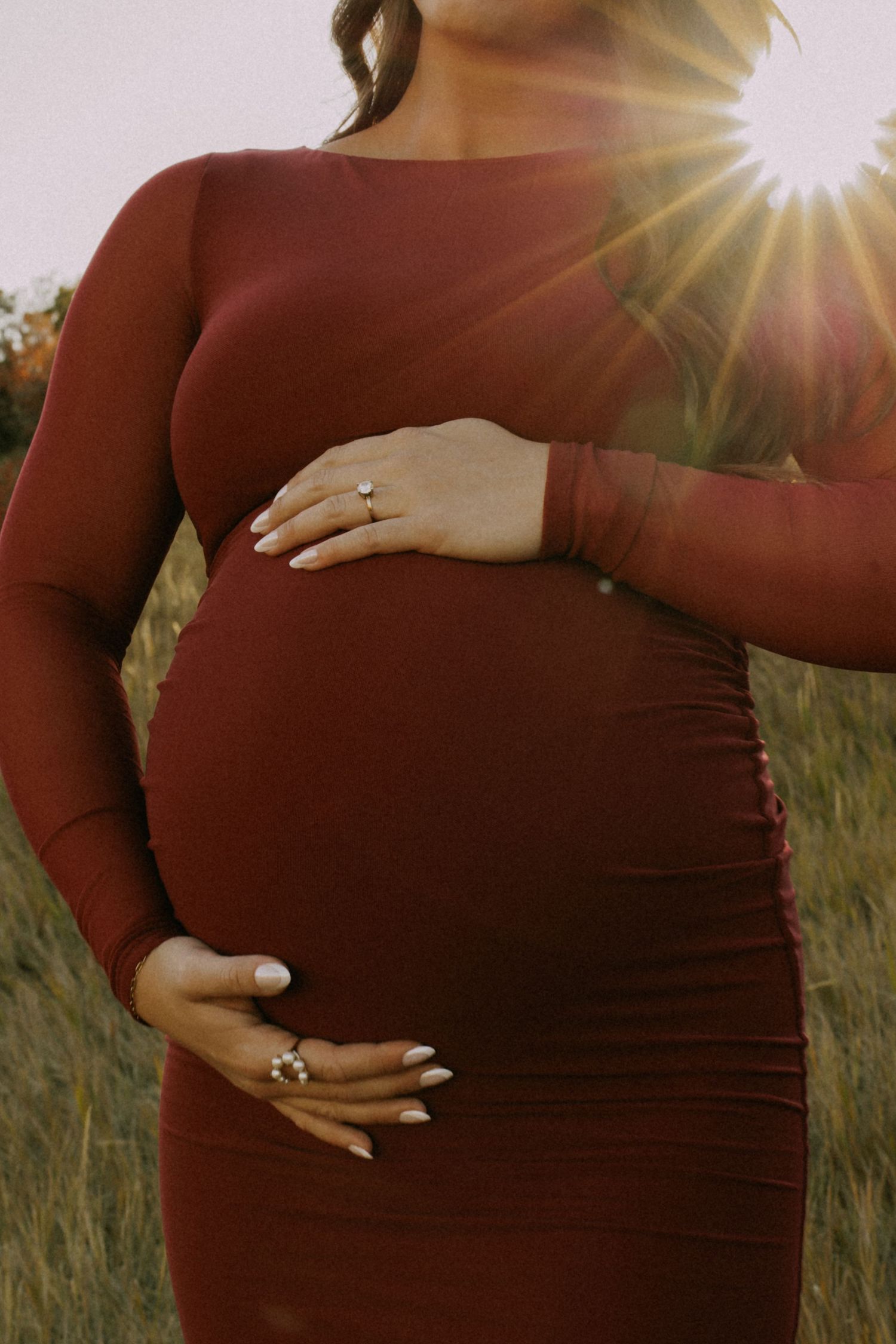 Maternity photo showing hands cradling baby bump in burgundy dress with sunlight flare.