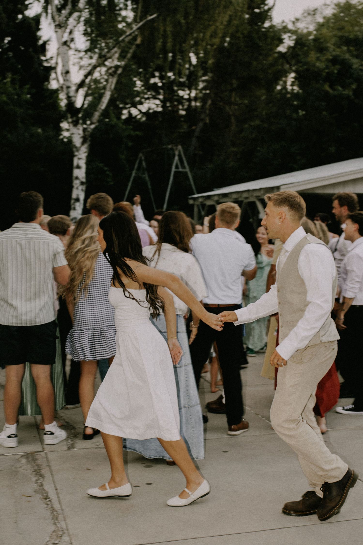 Group of wedding attendees dancing energetically outdoors in summer attire.
