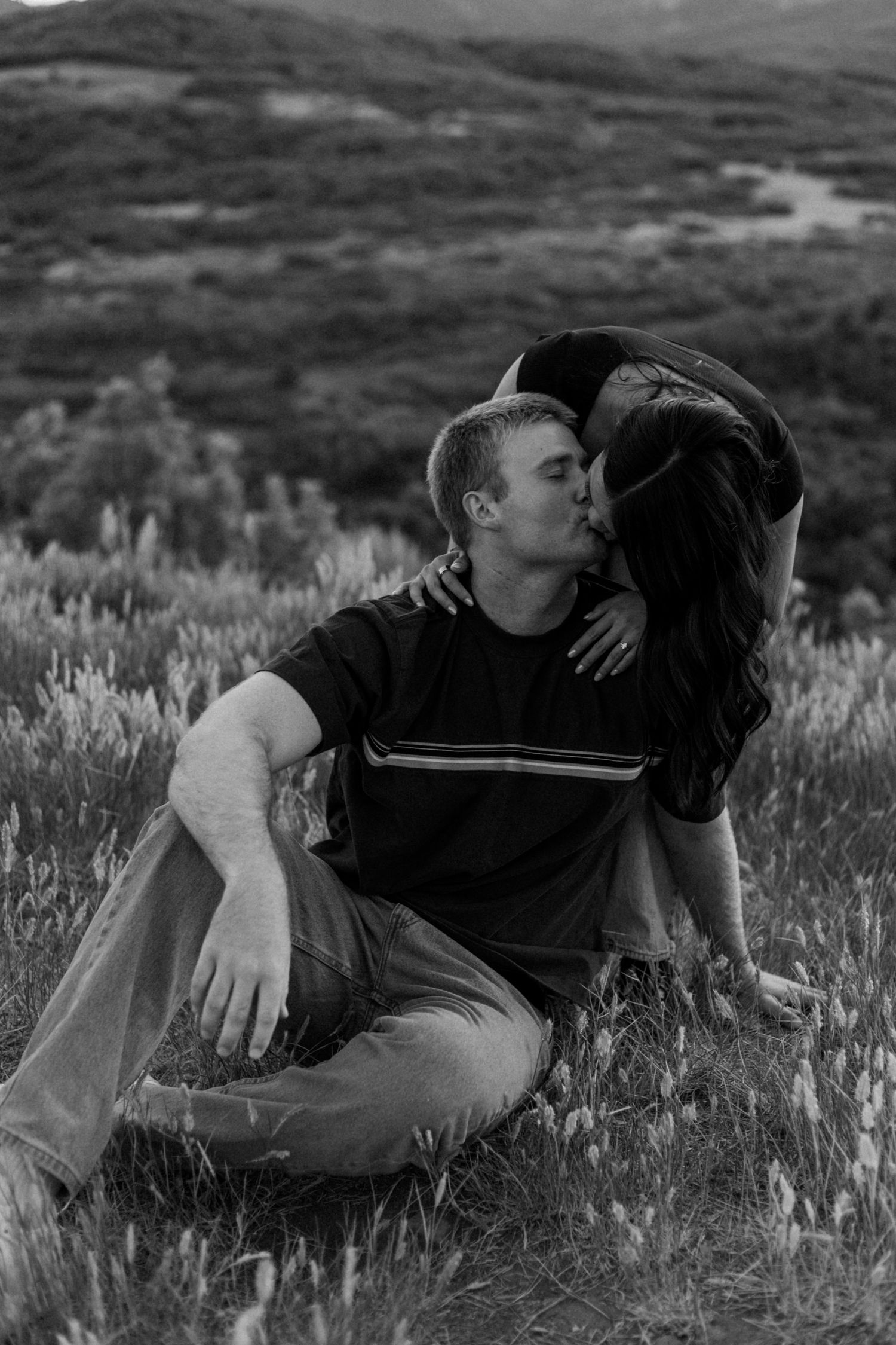 Two people share a tender moment while lying in a meadow with mountains in the distance.