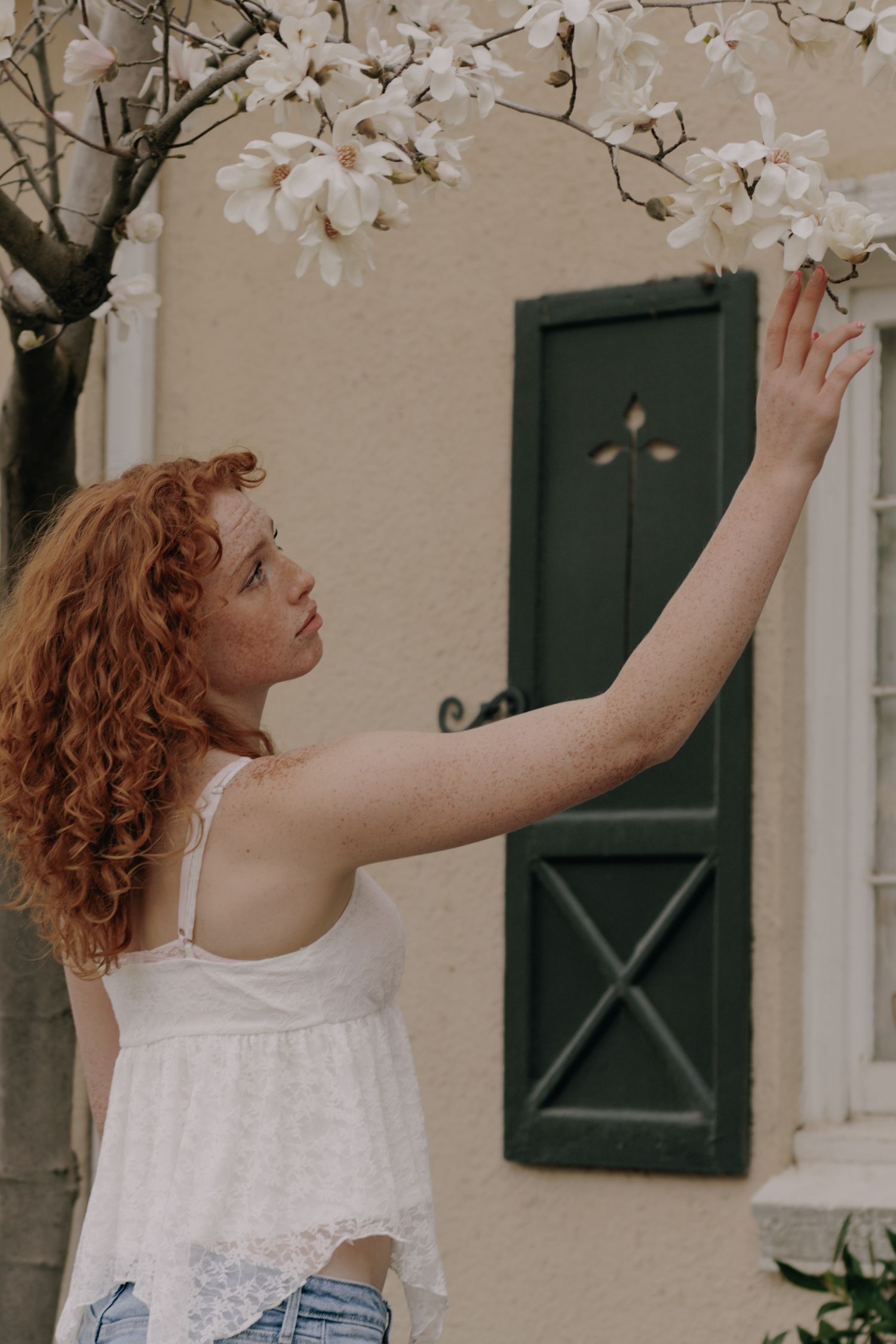 Someone in a white lace top reaching up to touch blooming spring tree branches against a beige wall.