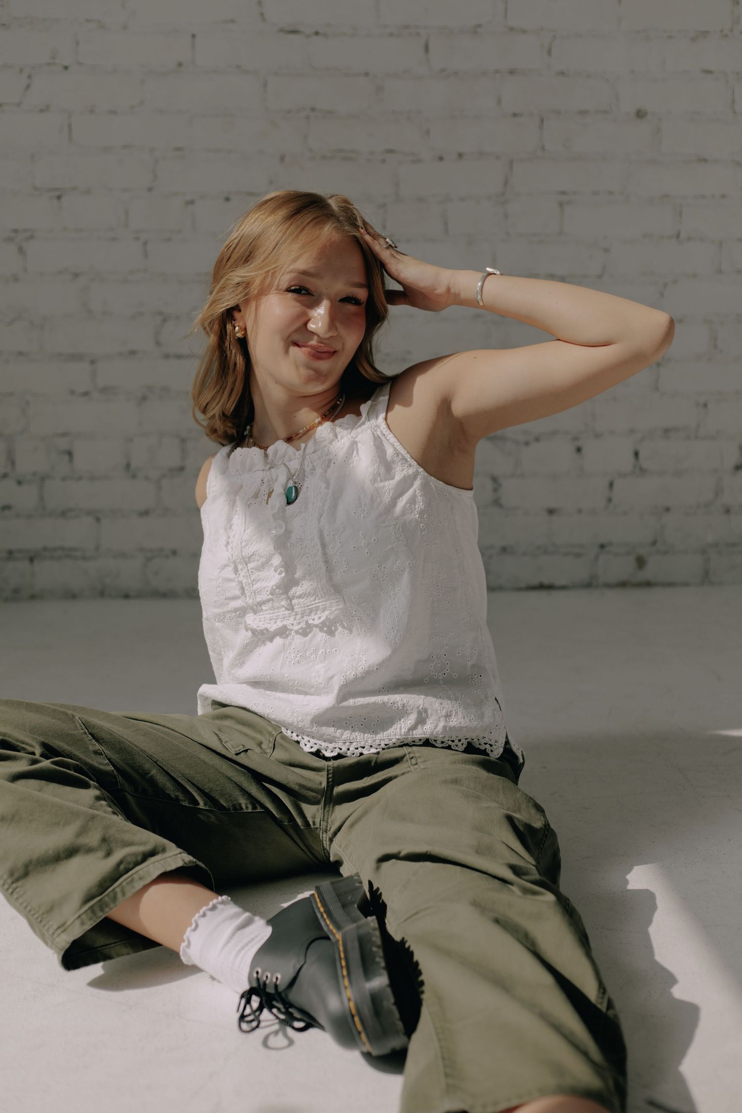 A person in a white top and olive pants poses playfully on a white floor against a brick wall.