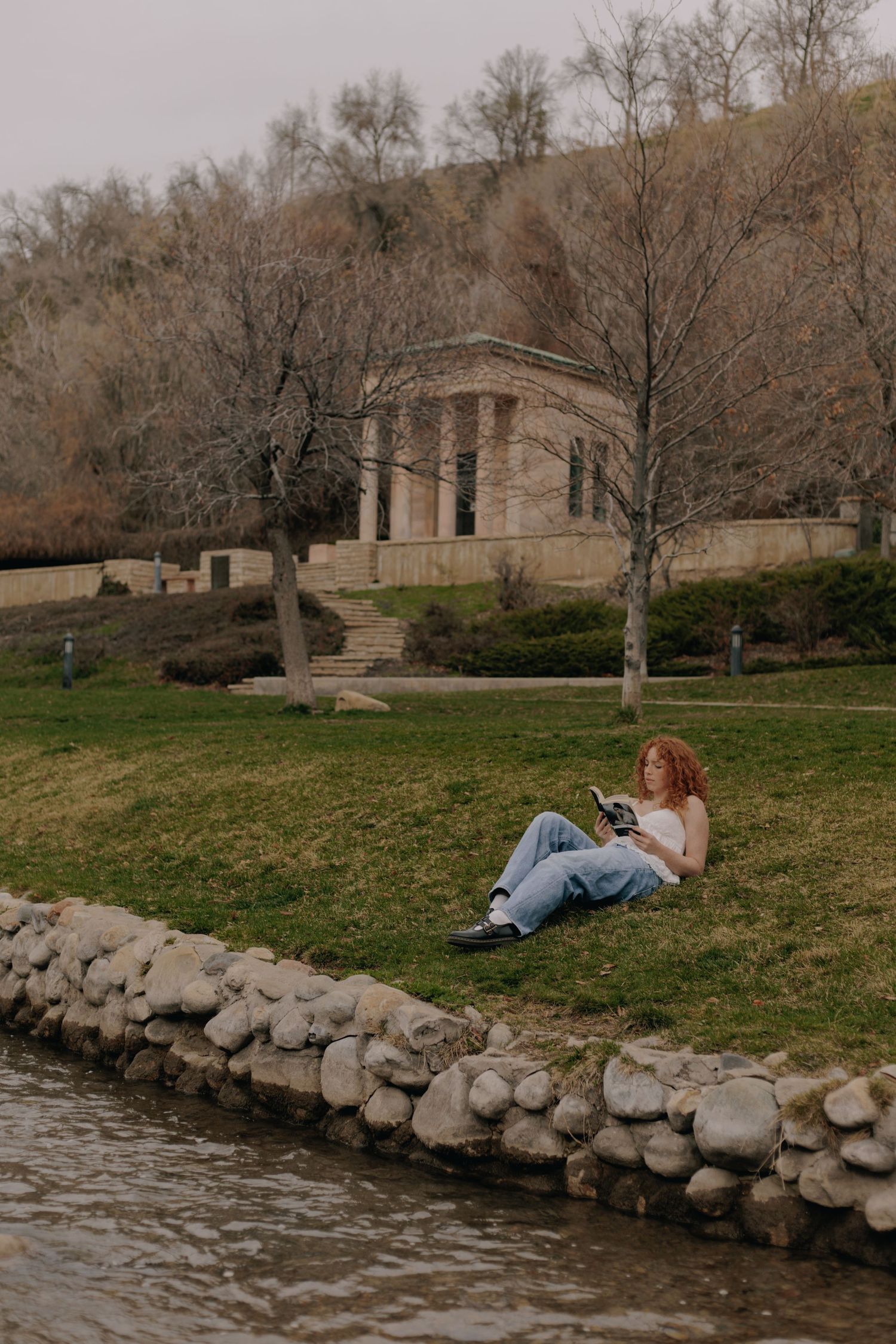 Person relaxing by stone-lined riverbank on grassy hill.