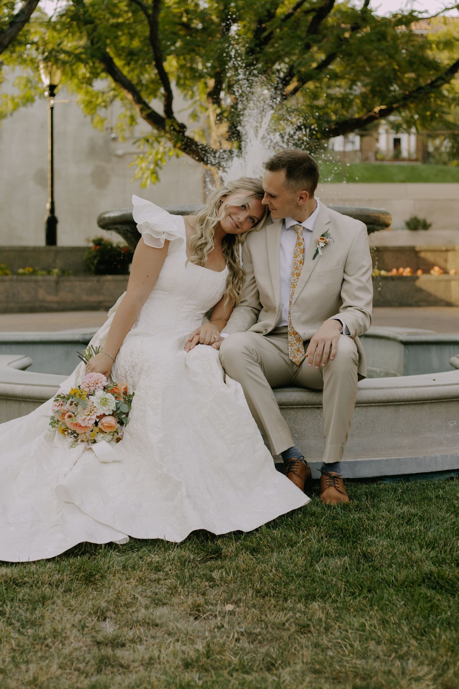 Couple in wedding attire sits together on lawn during golden hour photography session.