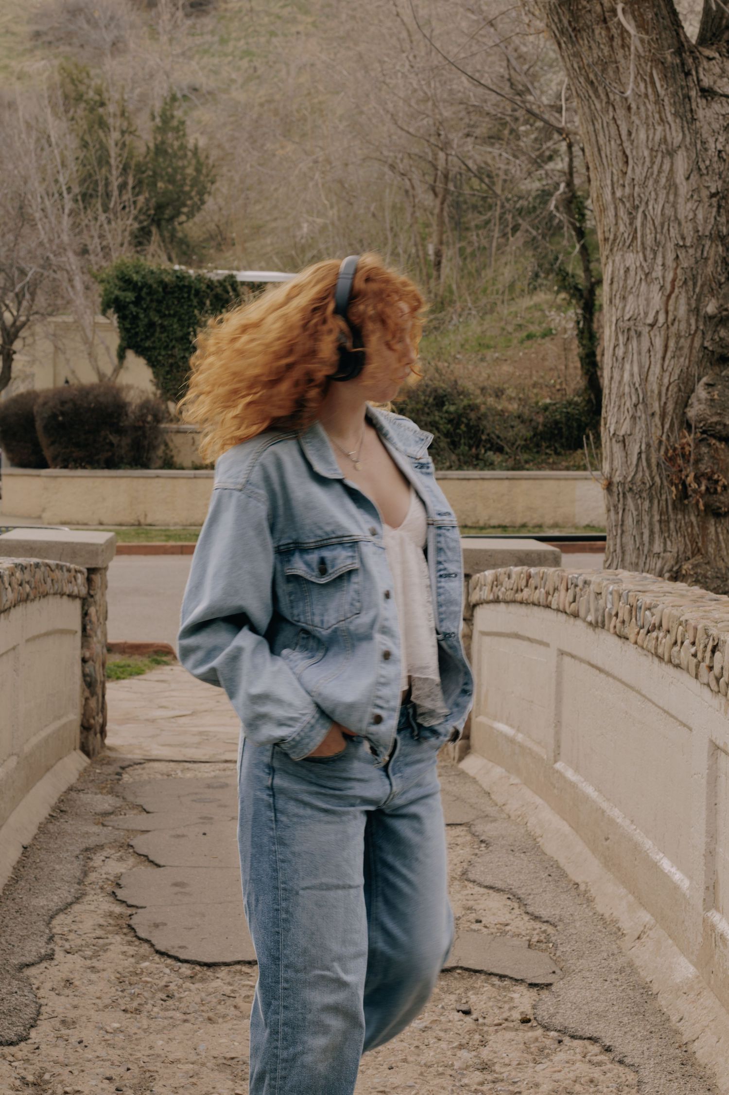 Person in denim outfit posing against curved stone wall outdoors.