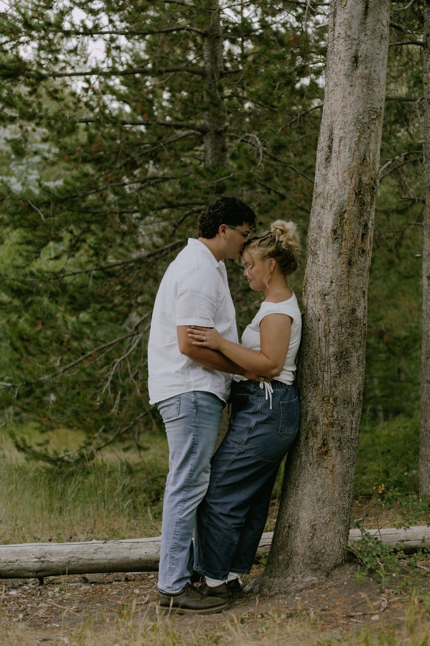 Two people share a romantic moment against a pine tree in a forest setting.