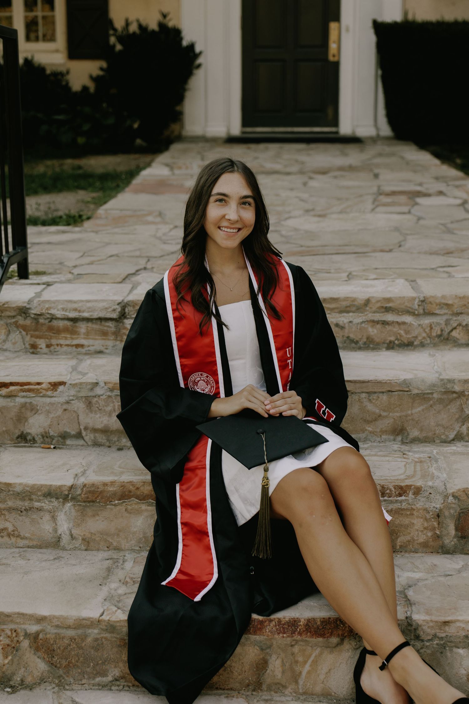 Graduate in cap and gown sits on stone steps during outdoor photo session.