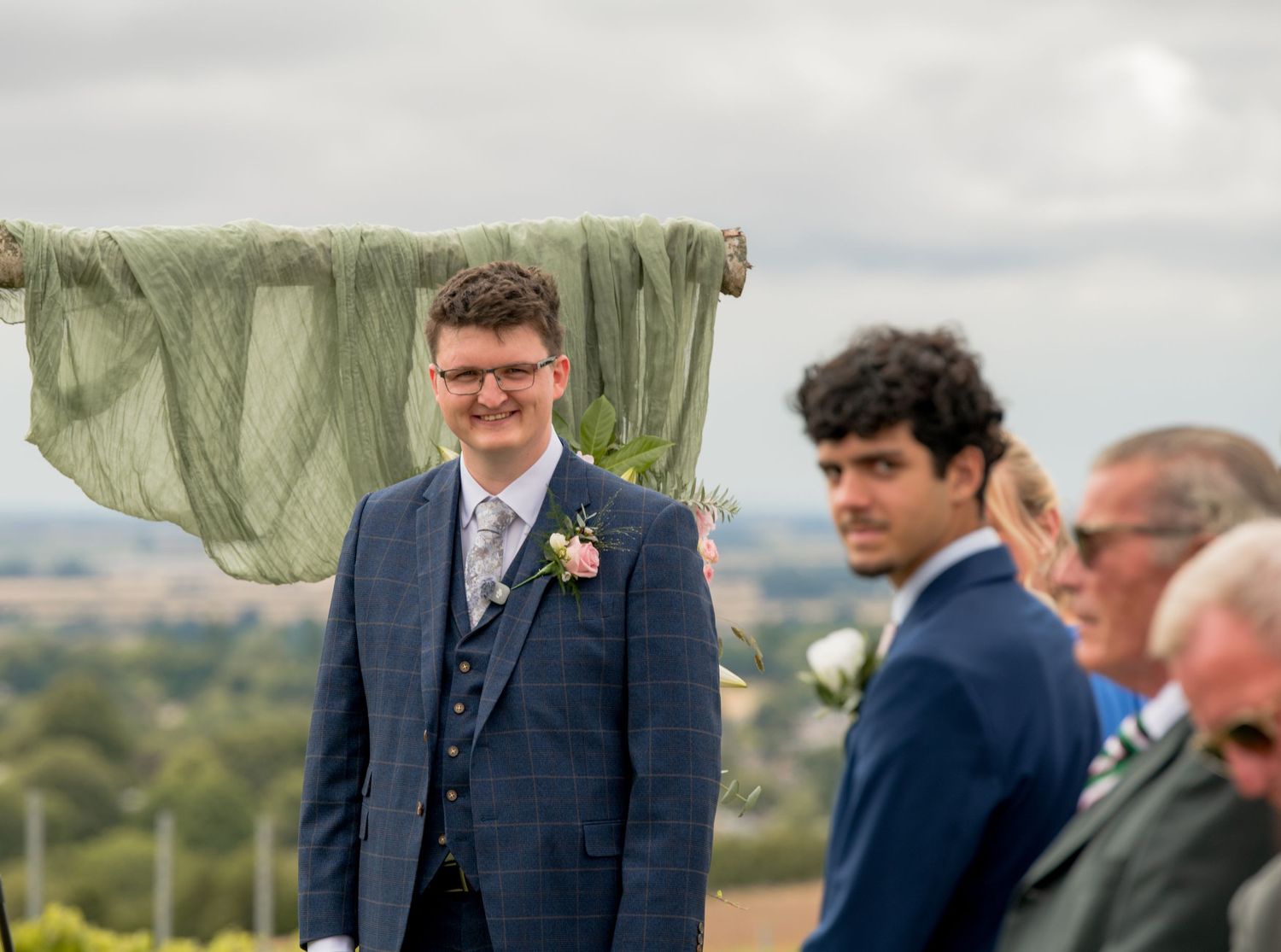 Smiling groom in navy suit with boutonniere awaiting bride at outdoor countryside wedding ceremony.