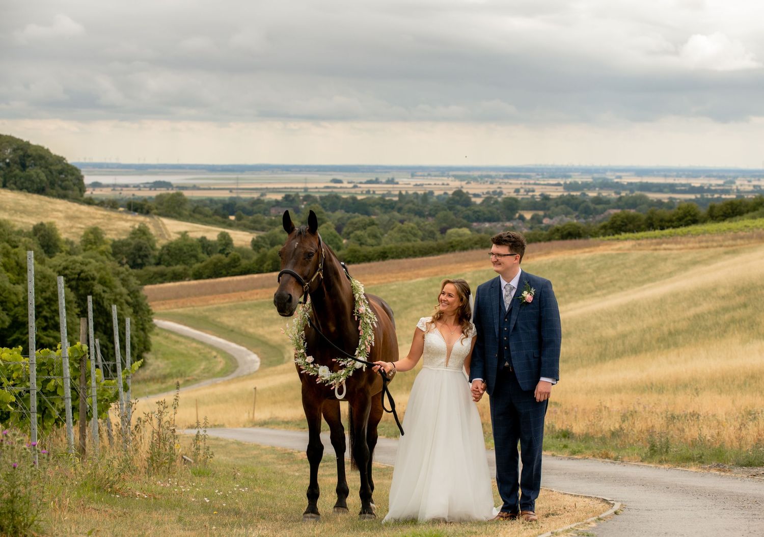 Bride and groom standing with dark horse on scenic vineyard road with rolling countryside views.