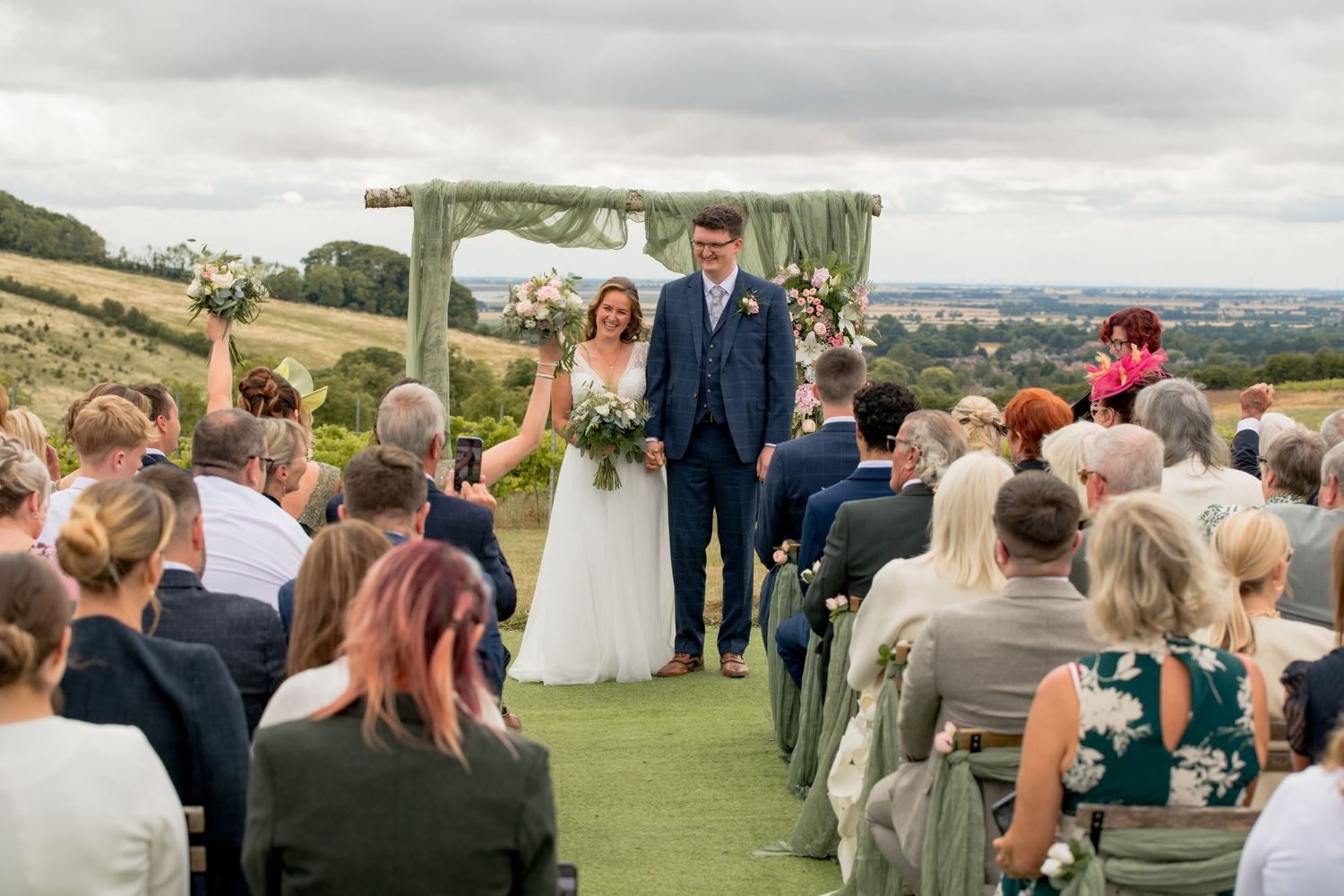 Outdoor wedding ceremony with bride and groom at floral arch altar overlooking rolling countryside.