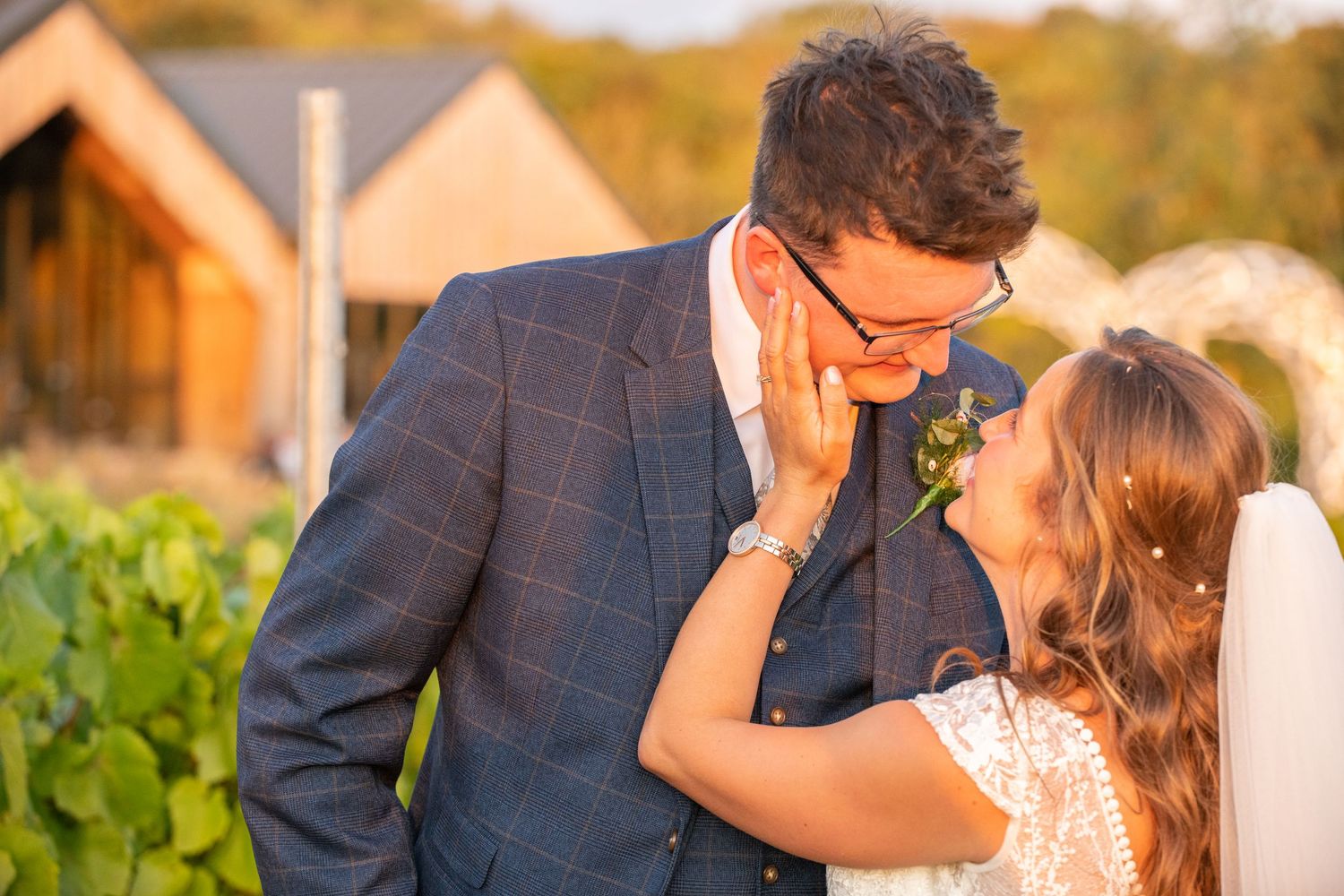 Groom in blue plaid suit tenderly kissing bride with pink boutonniere at golden hour.