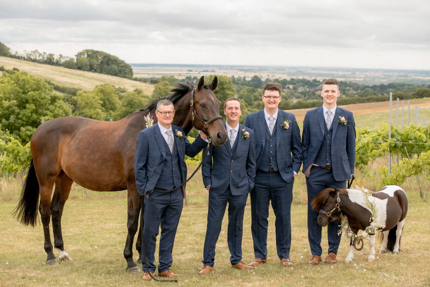 Three groomsmen in navy suits posing with brown horse and miniature pony against scenic countryside backdrop.