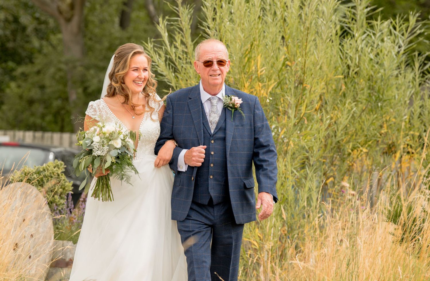 Bride in lace wedding dress walking down aisle with father through natural garden setting.