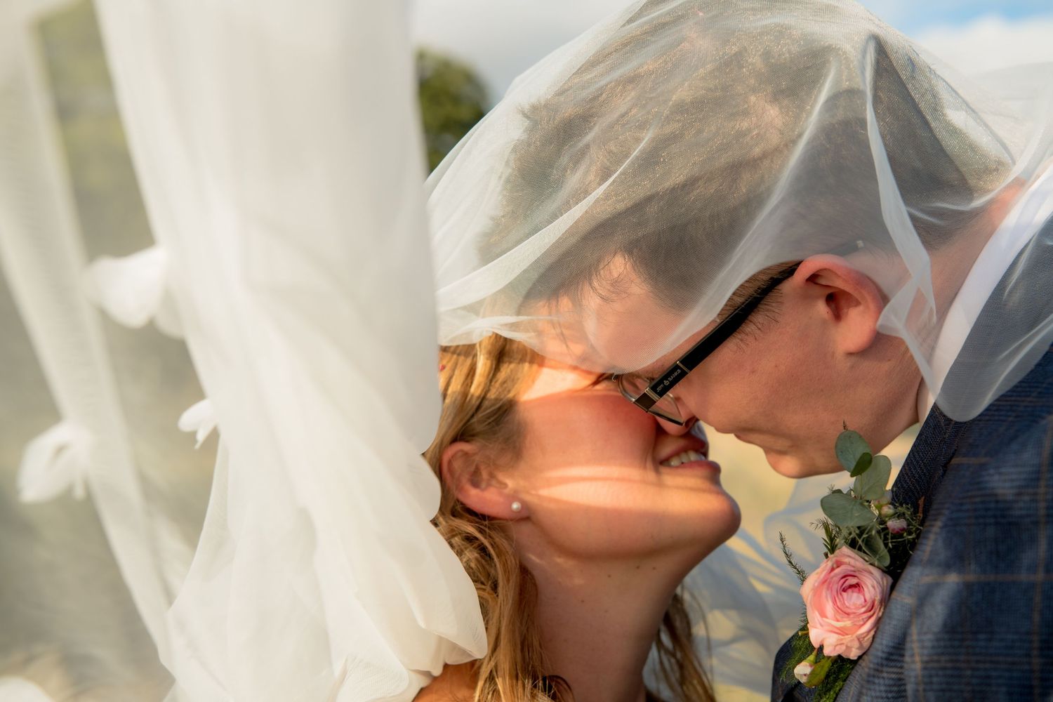 Bride and groom sharing passionate kiss under billowing white veil in bright sunlight.