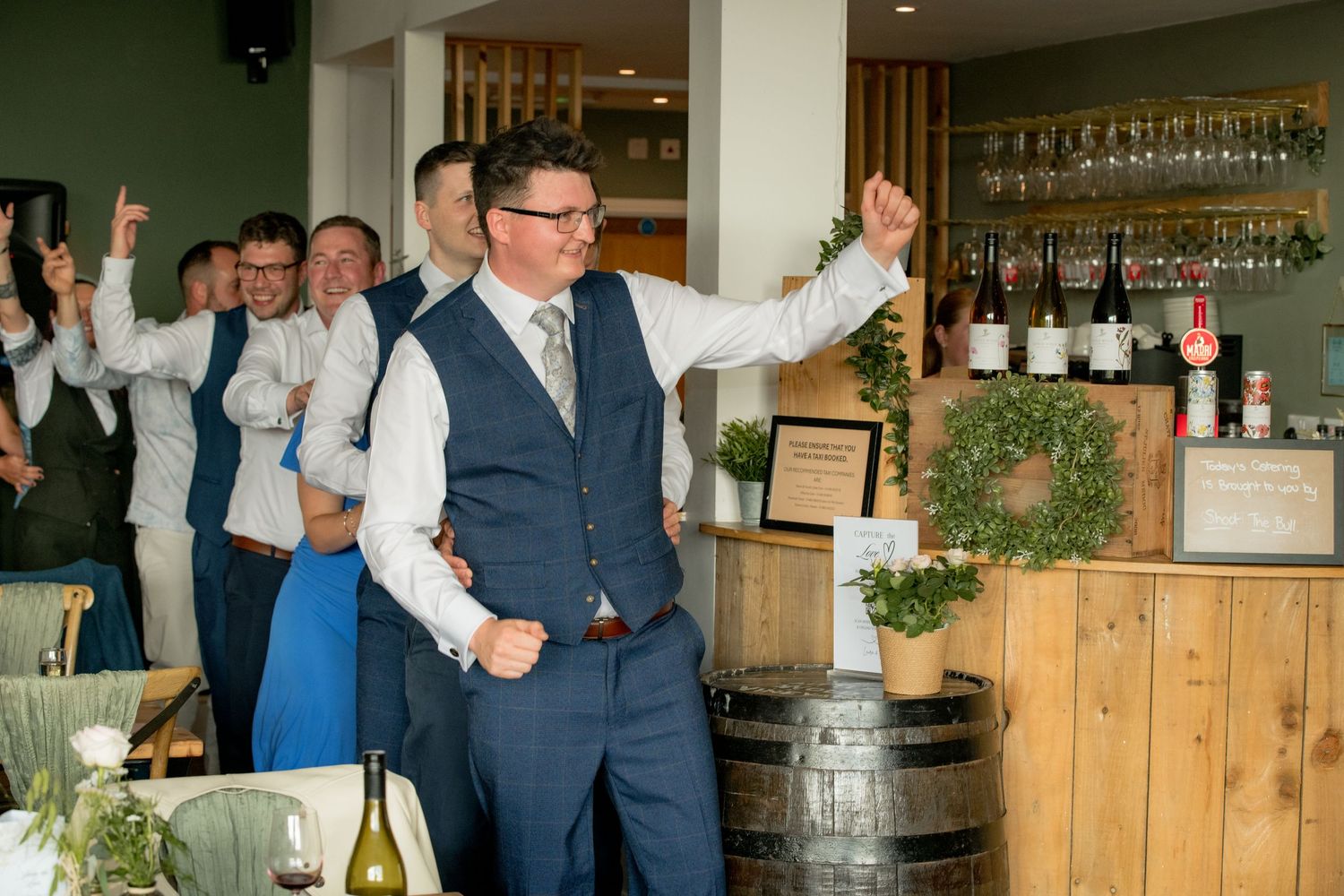 Groom celebrating with groomsmen raising hands in a rustic vineyard bar venue during wedding reception.