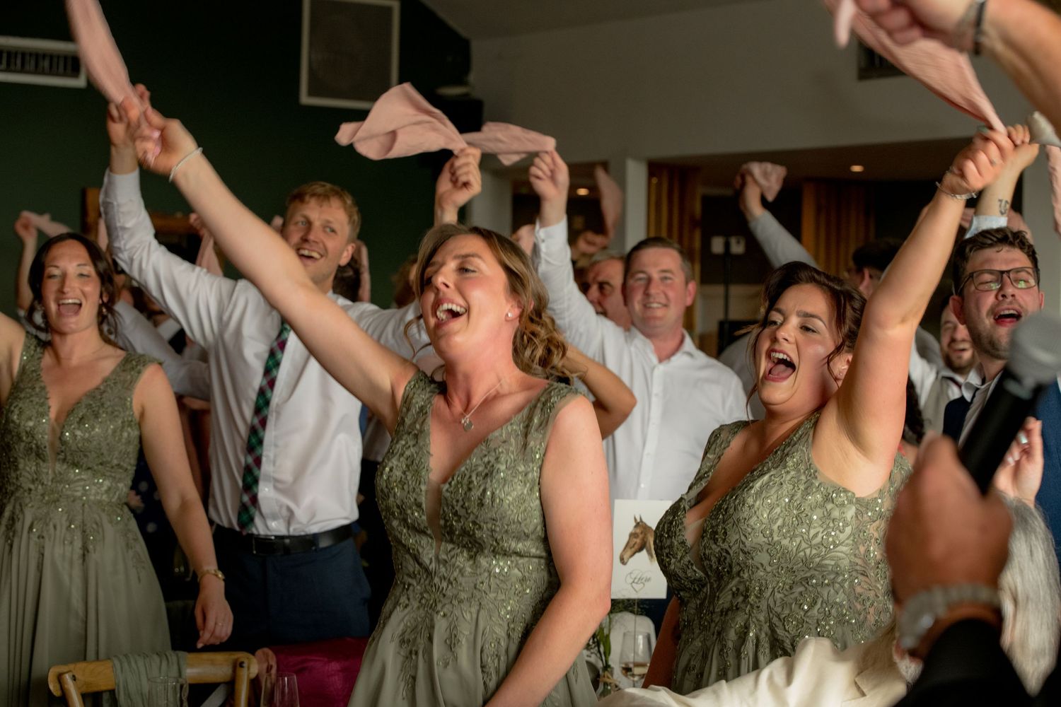 Bridesmaids in sage green dresses celebrating joyfully with raised arms at lively wedding reception.