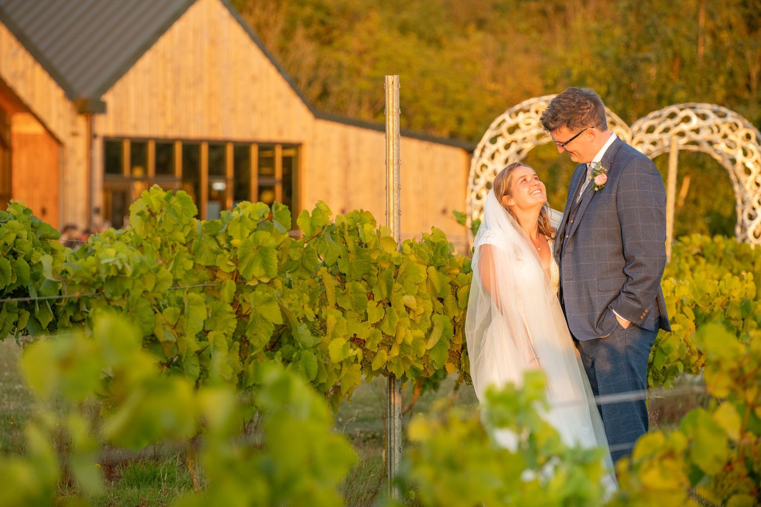 Bride and groom sharing tender moment in vineyard with rustic barn and horse shoe arch backdrop.