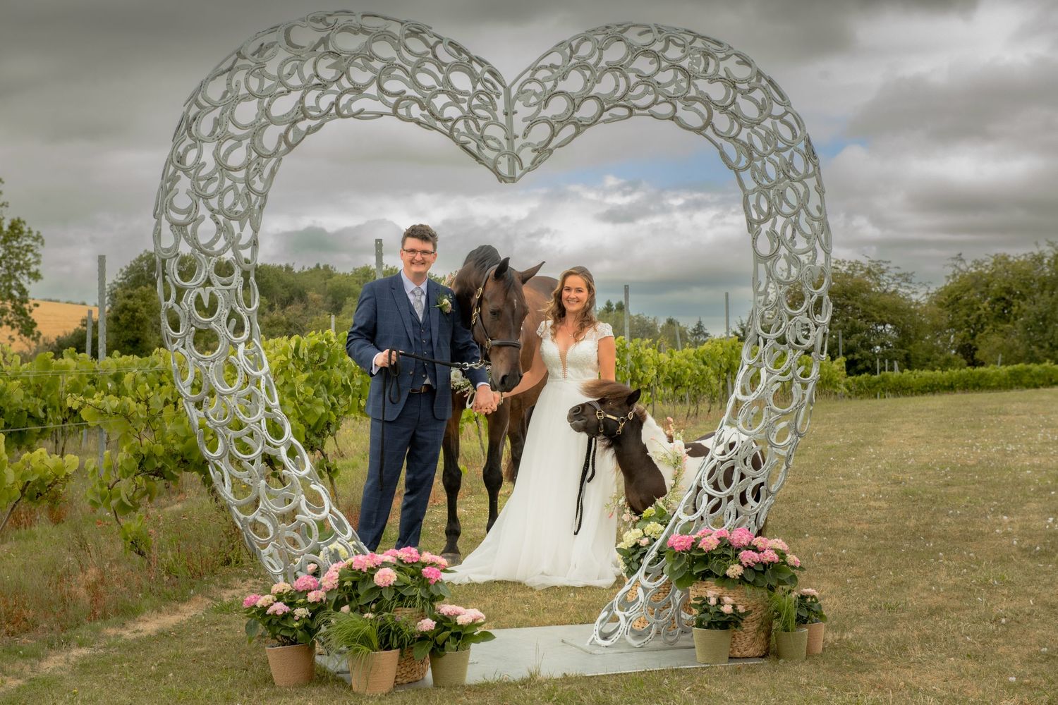 Bride and groom posing with dark horse under ornate heart-shaped horse shoe arch at vineyard wedding ceremony.