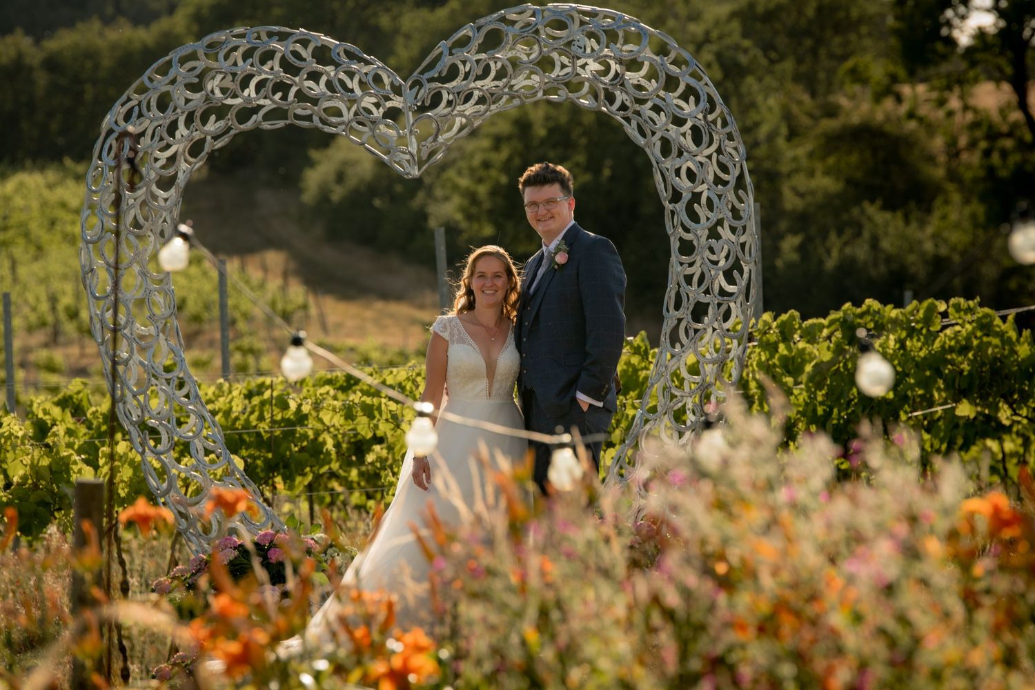 Bride and groom standing together under horseshoe heart arch surrounded by colorful flowers in vineyard at golden hour.