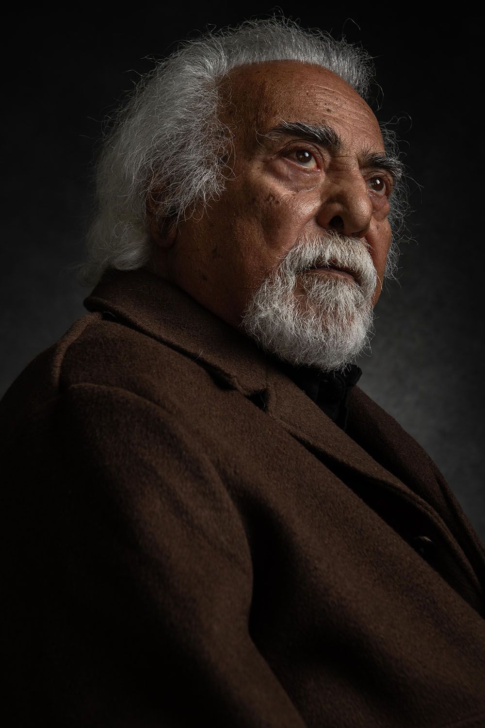 Elderly man with white hair and beard wearing a brown coat, gazing thoughtfully against a dark background.
