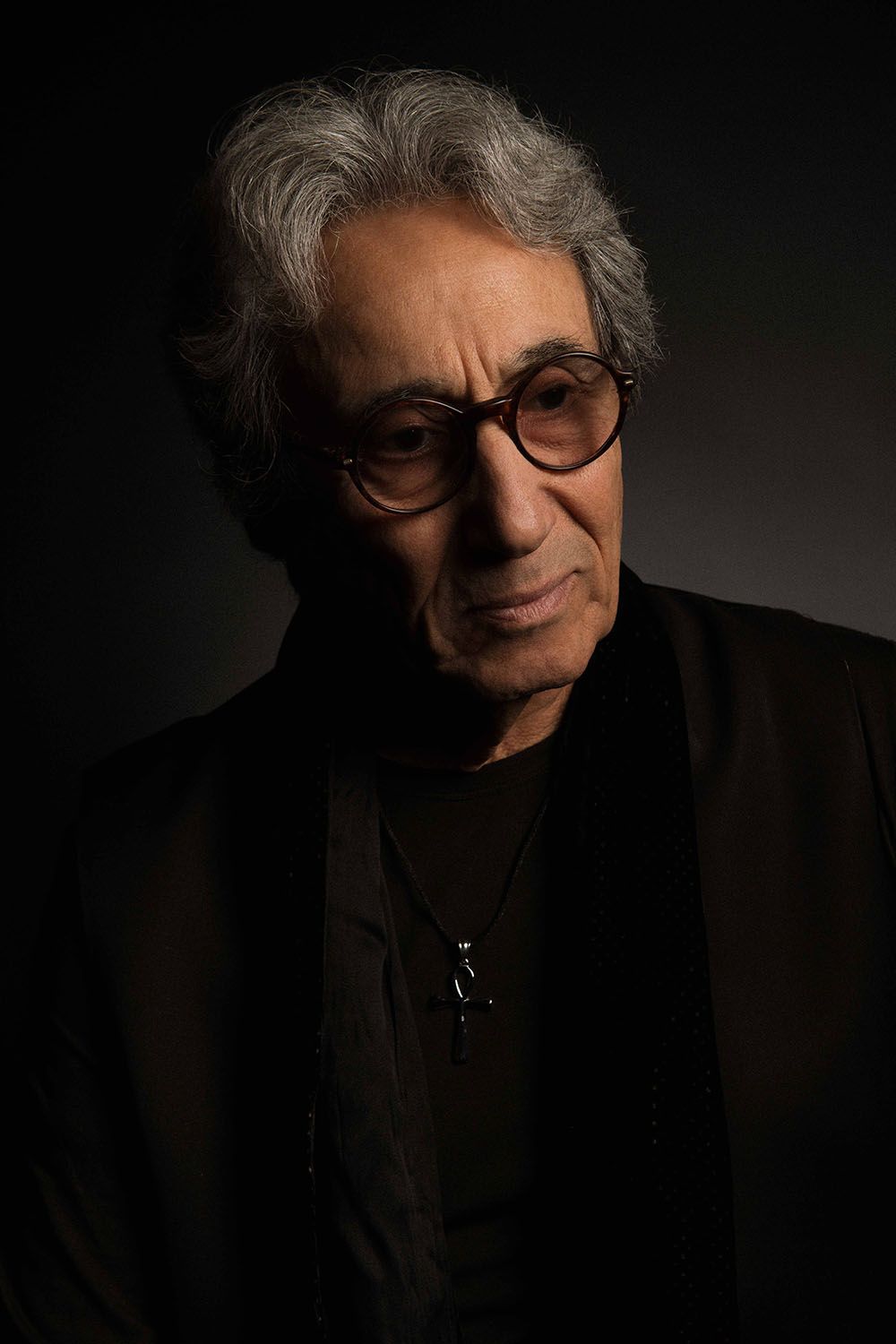 Older man with gray hair and round glasses wearing a black jacket, dramatic studio portrait with dark background.