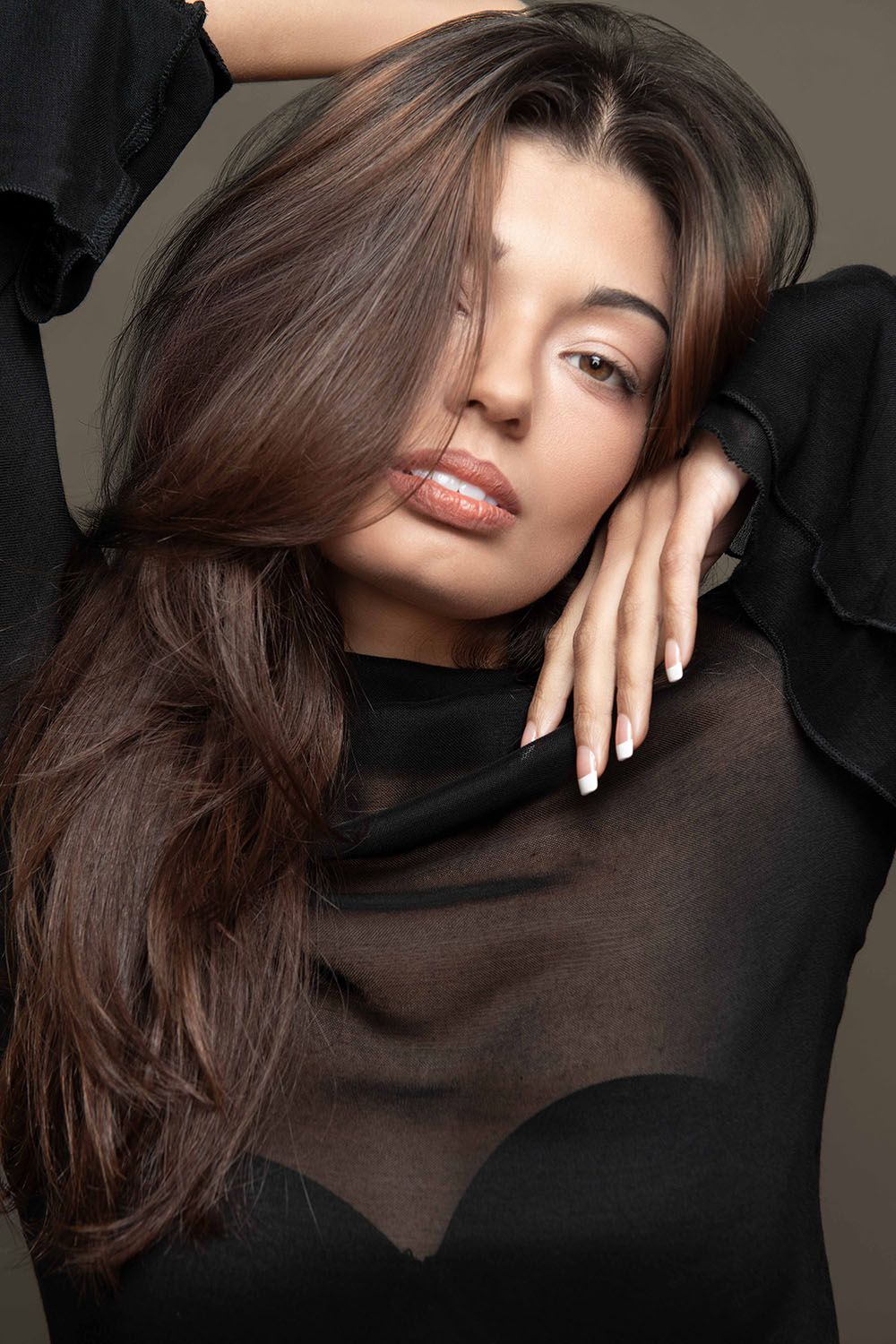 Brunette woman in black sheer top poses with hair falling over face, hands crossed at chest, studio beauty shot.
