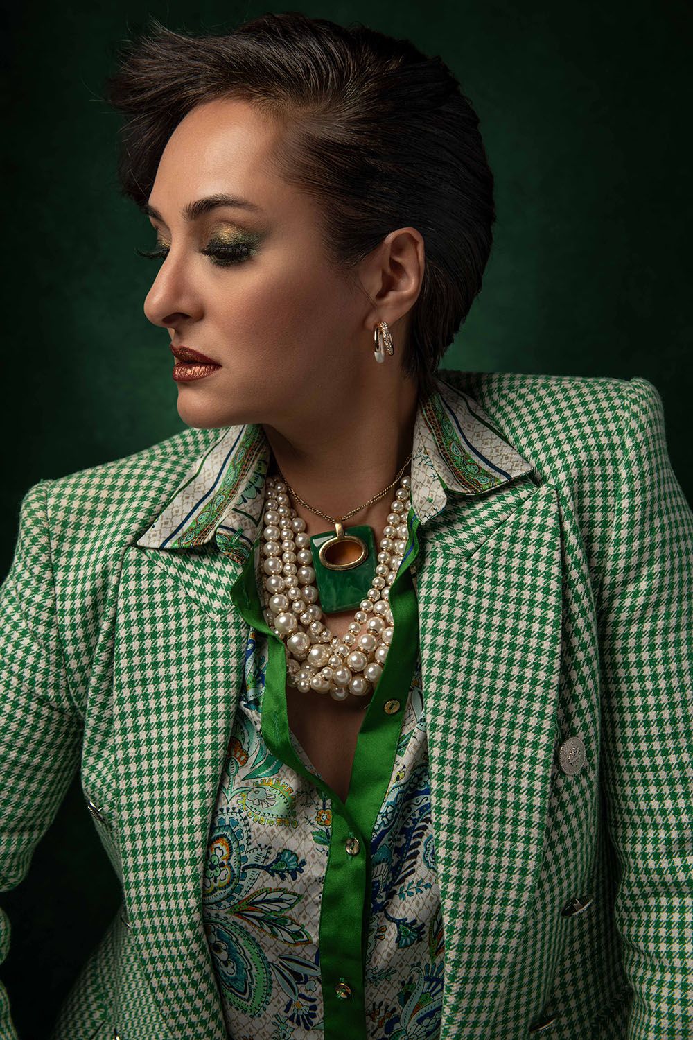 Woman in green houndstooth blazer with pearl necklace and floral blouse poses against dark green background.