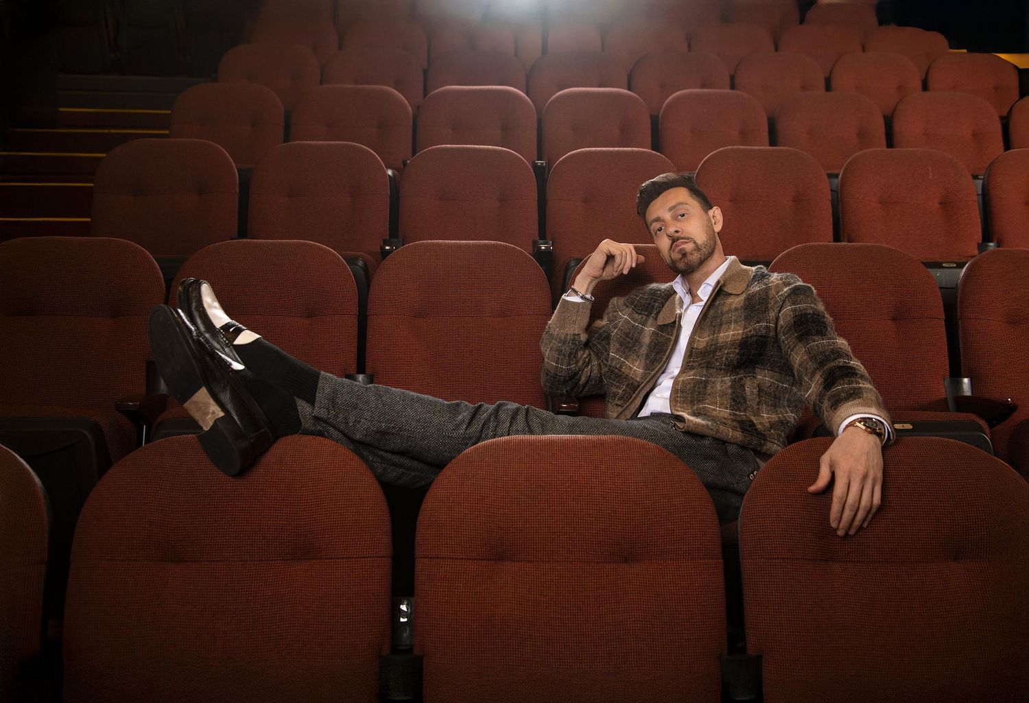 portrait of iranian american comedian Max Amini lounging casually across red kia forum seats in cinematic lighting.