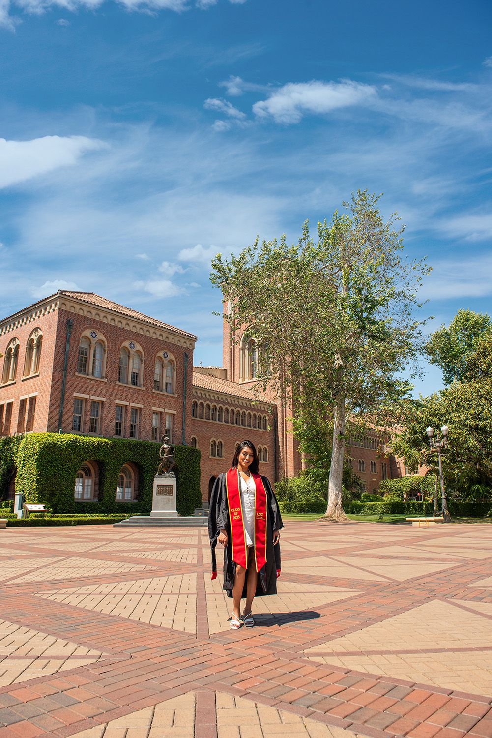 Graduate in cap and gown with red stole poses on brick plaza before historic red brick university building under blue sky.