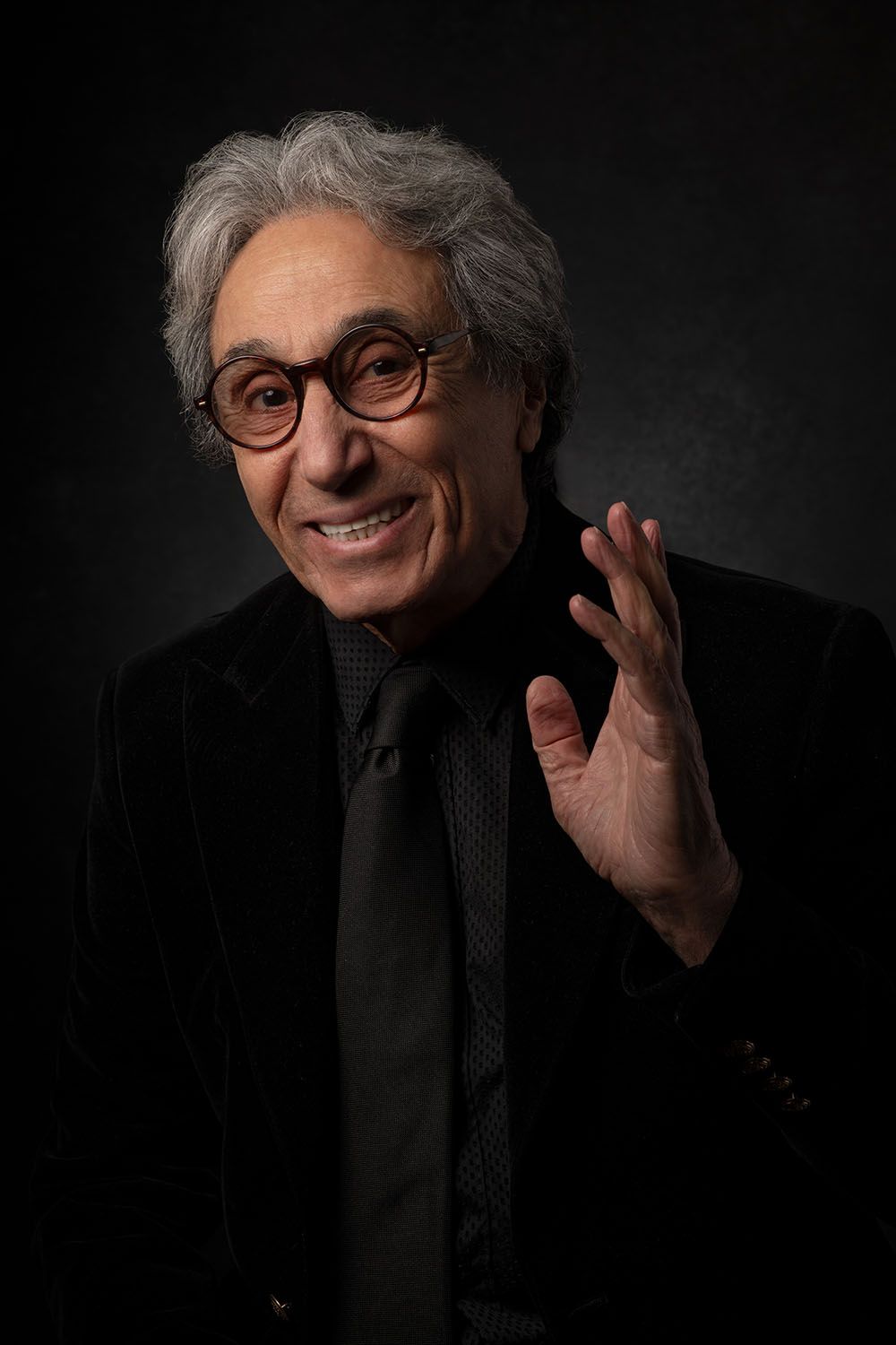 Smiling elderly man with gray hair, round glasses, black suit and tie, gesturing with hand against dark background.