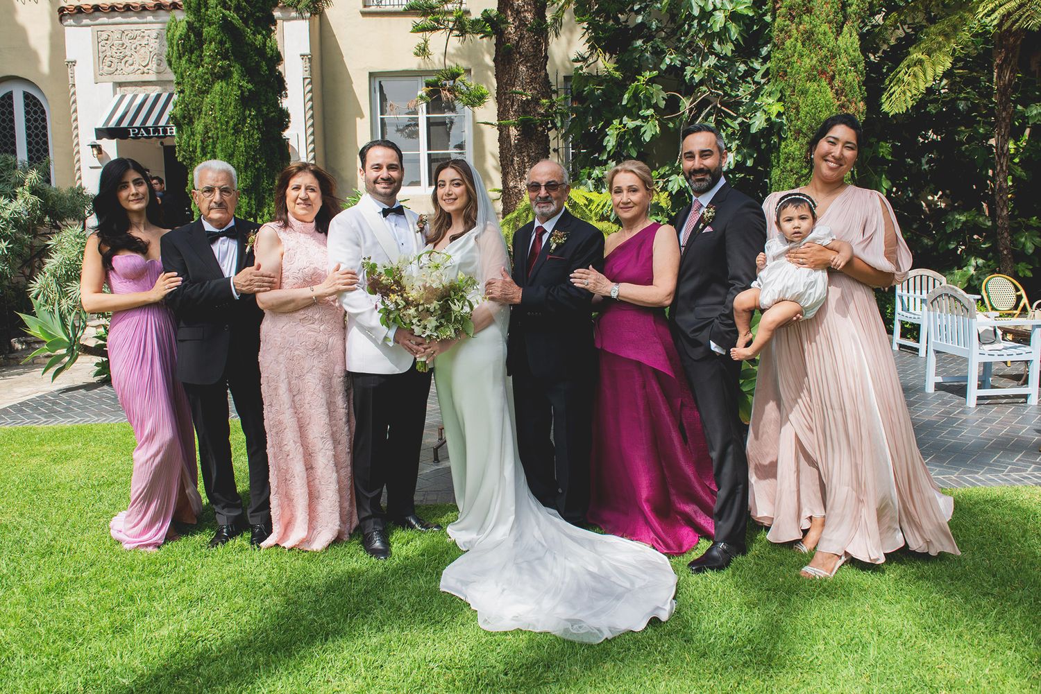 Wedding party members in pastel pink and burgundy dresses pose together in an outdoor garden setting.