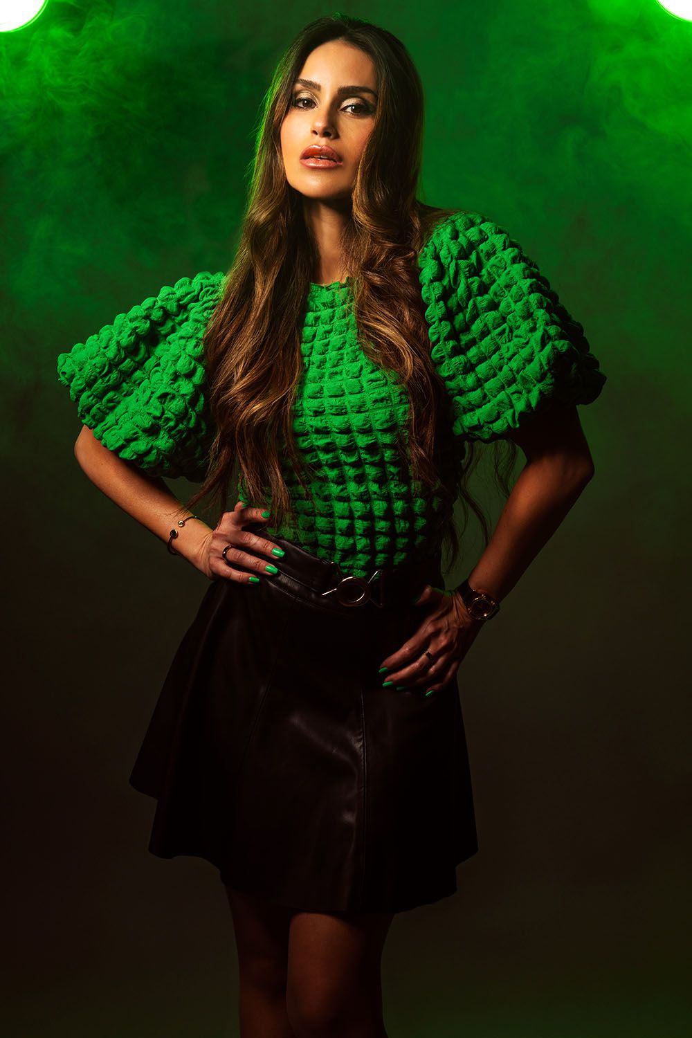 Woman in textured green puff-sleeve top and black leather mini skirt posing against dark background with green lighting.
