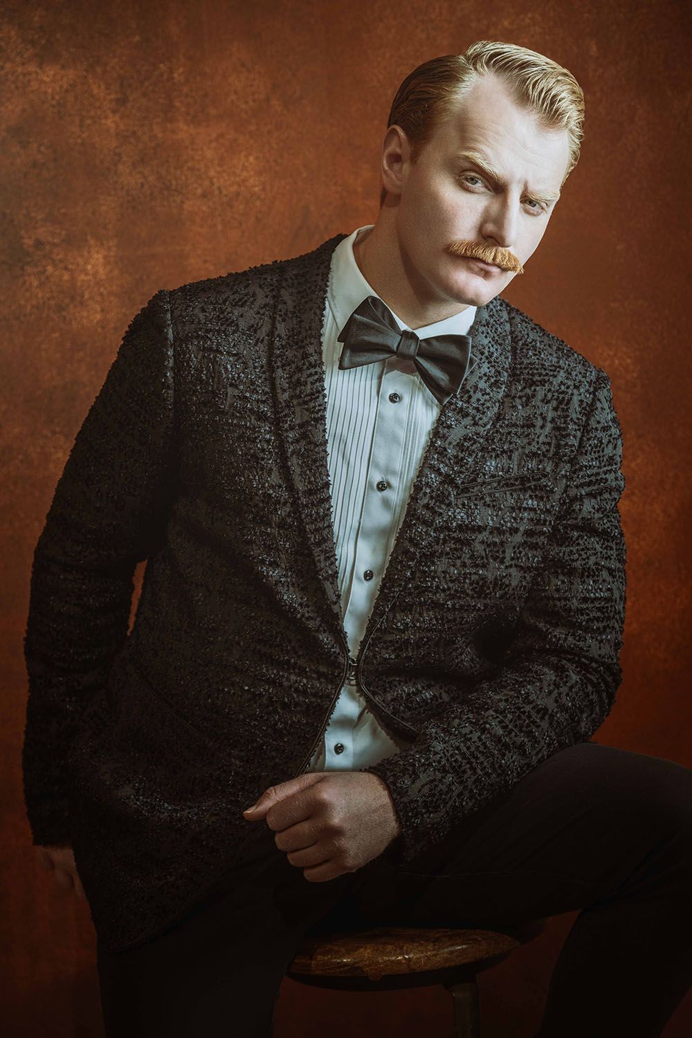 Stylish blond man with mustache wearing dark tweed blazer and bow tie sits on stool against rustic brown background.