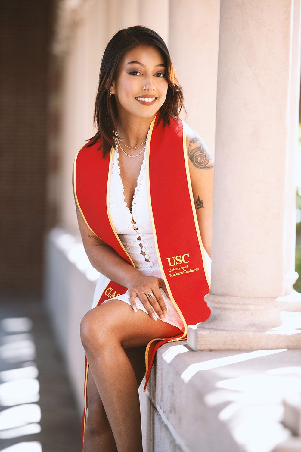 USC graduate in red and gold stole poses confidently between white columns in a white dress.